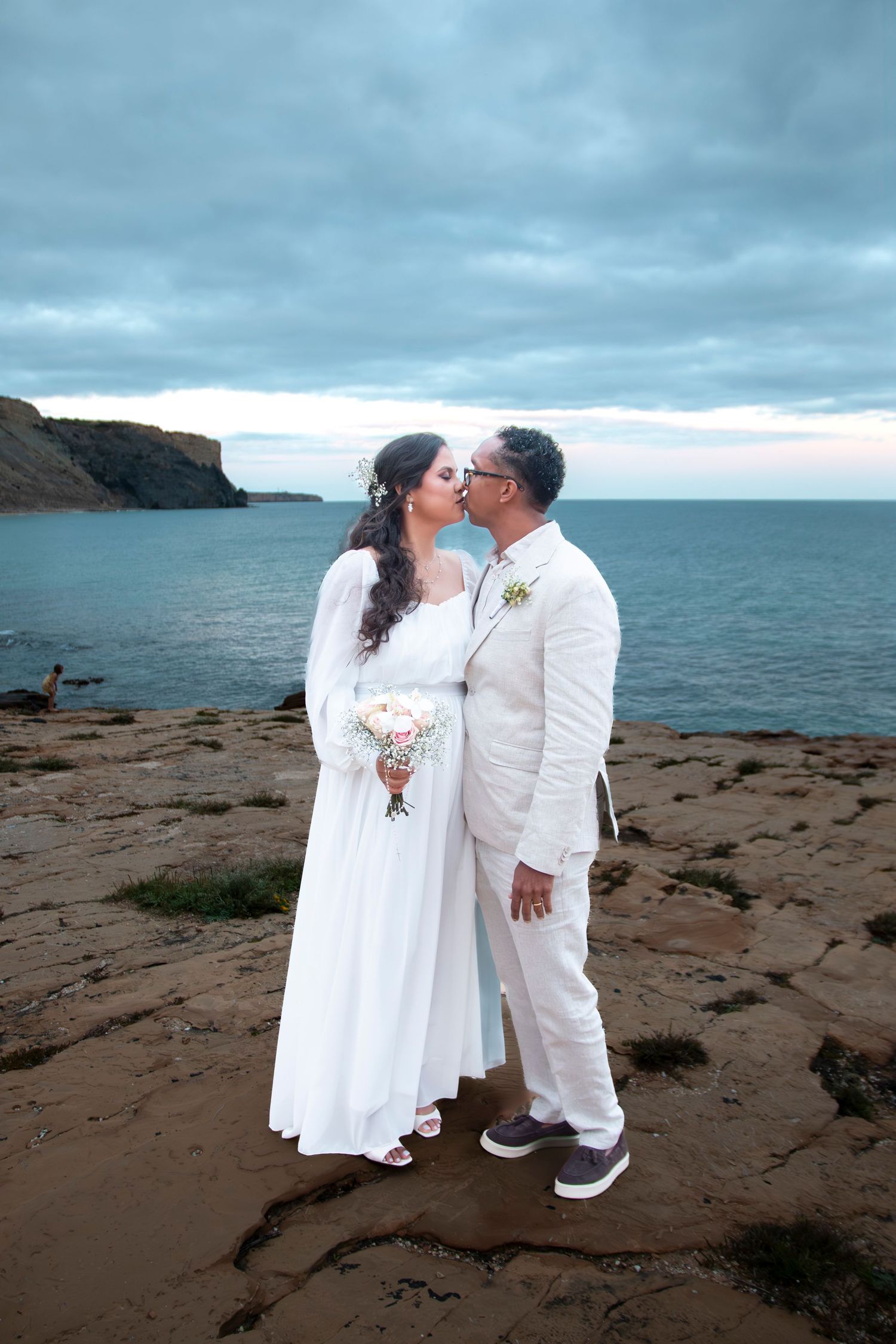 A couple in white wedding attire share an intimate moment on a rocky coastal beach under moody gray skies.