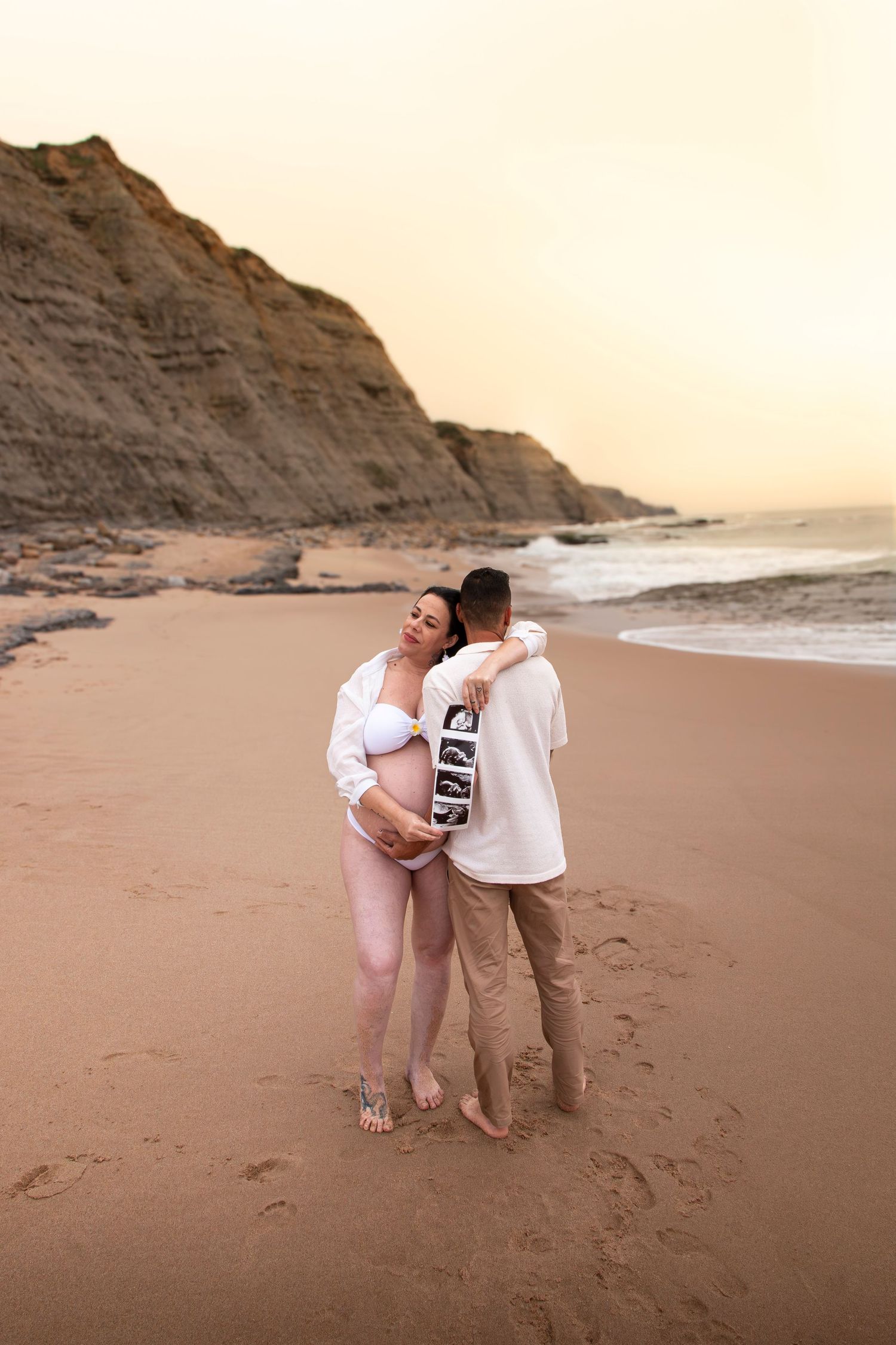 Pregnant couple embracing on a sandy beach at sunset, holding ultrasound photos, with cliffs and ocean waves in the background.