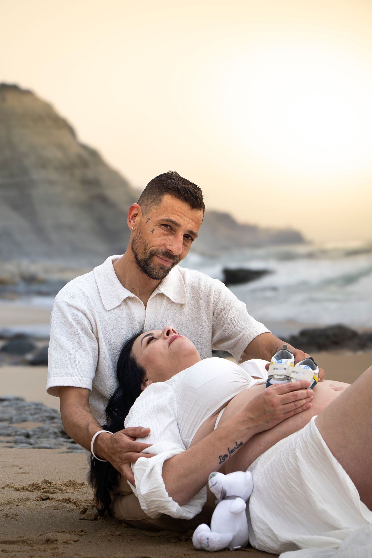Pregnant couple sitting on beach at sunset, woman lying back holding baby shoes on her belly, man gazing lovingly at her.