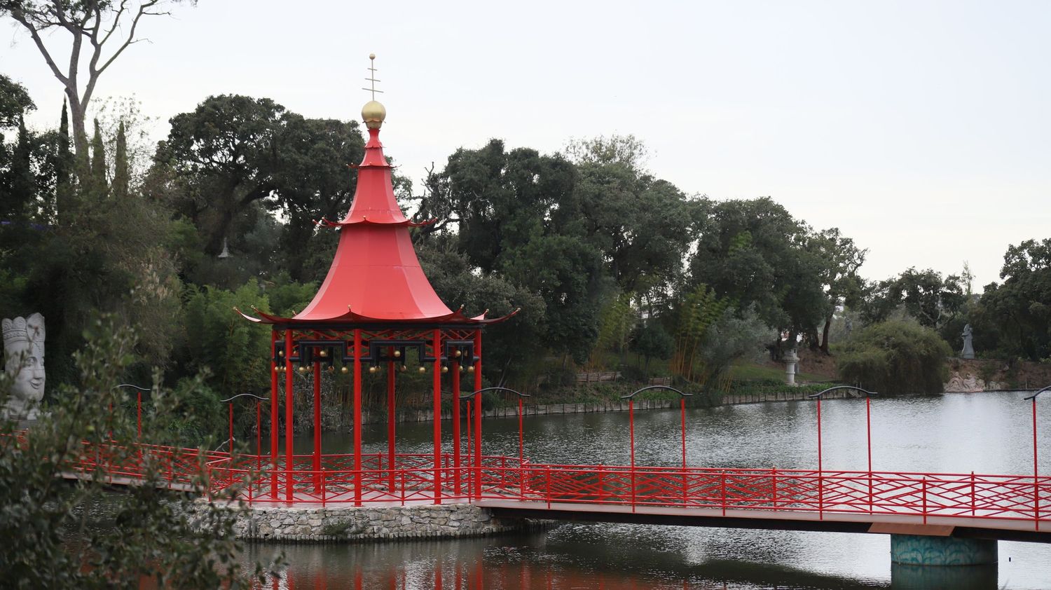 A scenic panoramic view of a red Asian-style pagoda and bridge over a tranquil lake in a lush garden setting.