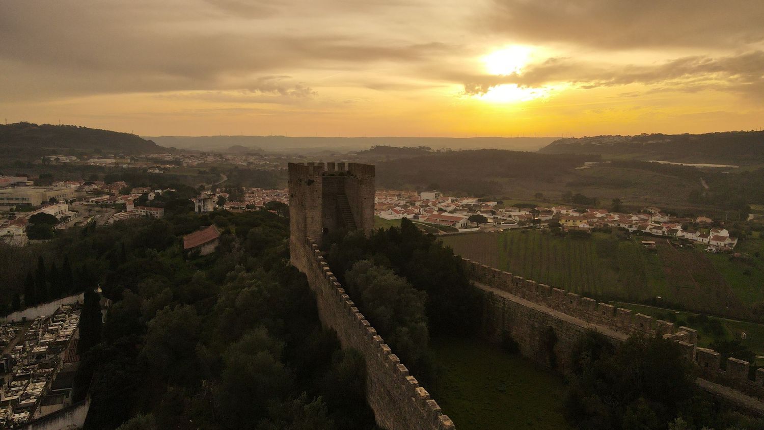 Ancient stone fortress wall stretches along hilltop at sunset with golden sky and rural village below in Portugal.