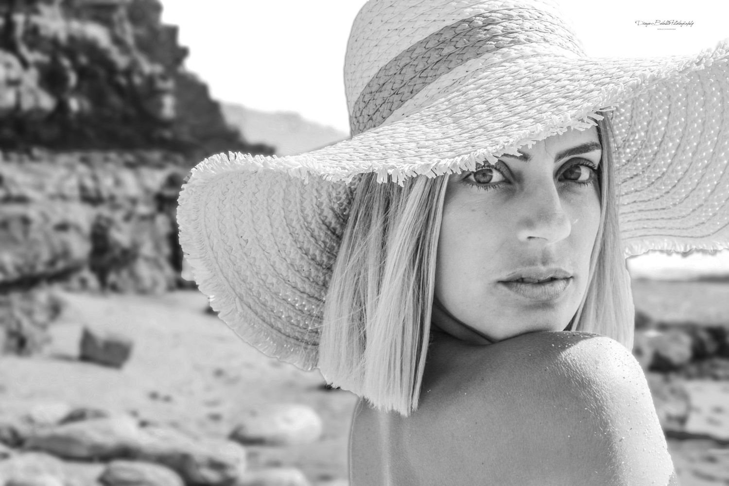 Black and white closeup photo of someone wearing a wide brimmed summer hat with blonde hair at the beach.