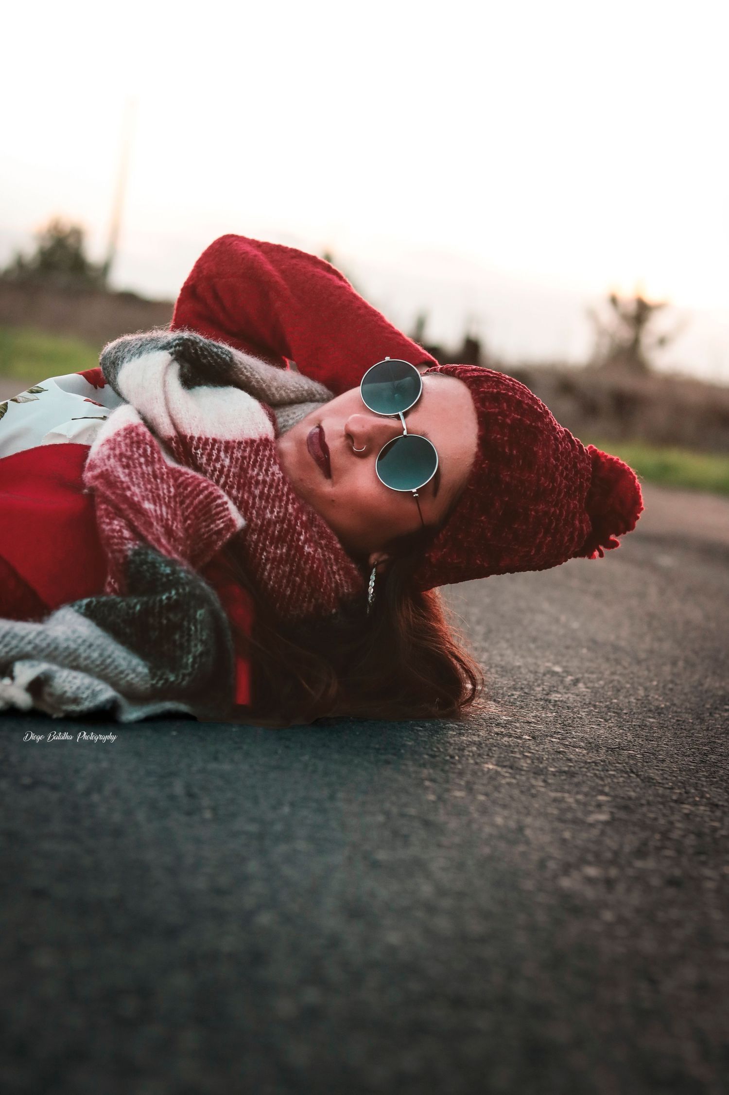 Person lying on ground wearing red knit hat, round sunglasses and winter clothing in artistic outdoor portrait.
