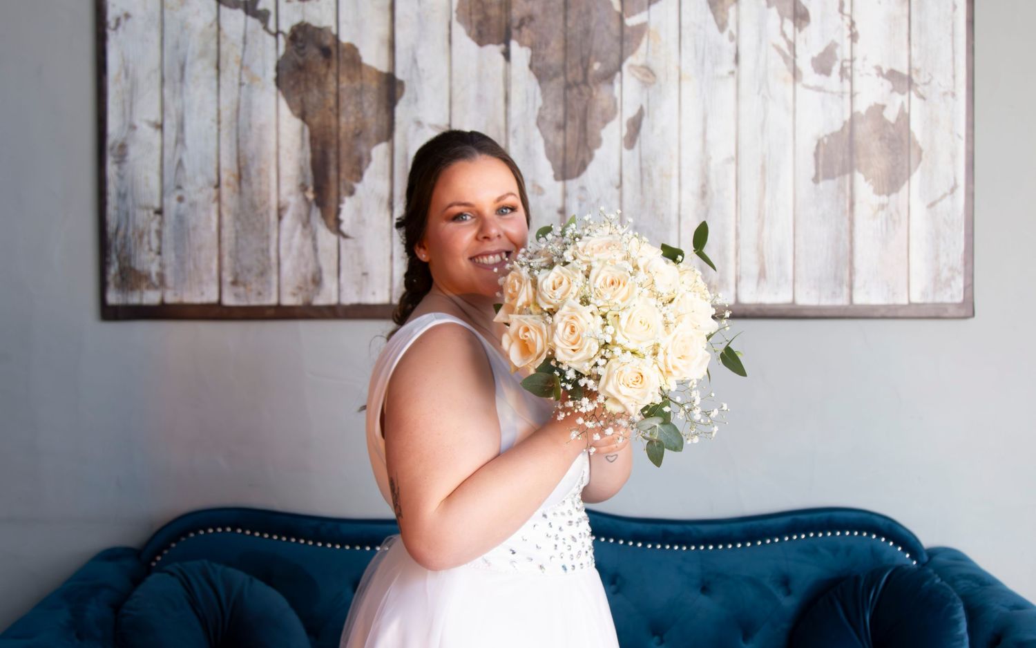 A bride poses with her white floral bouquet against a blue couch and wooden wall art.