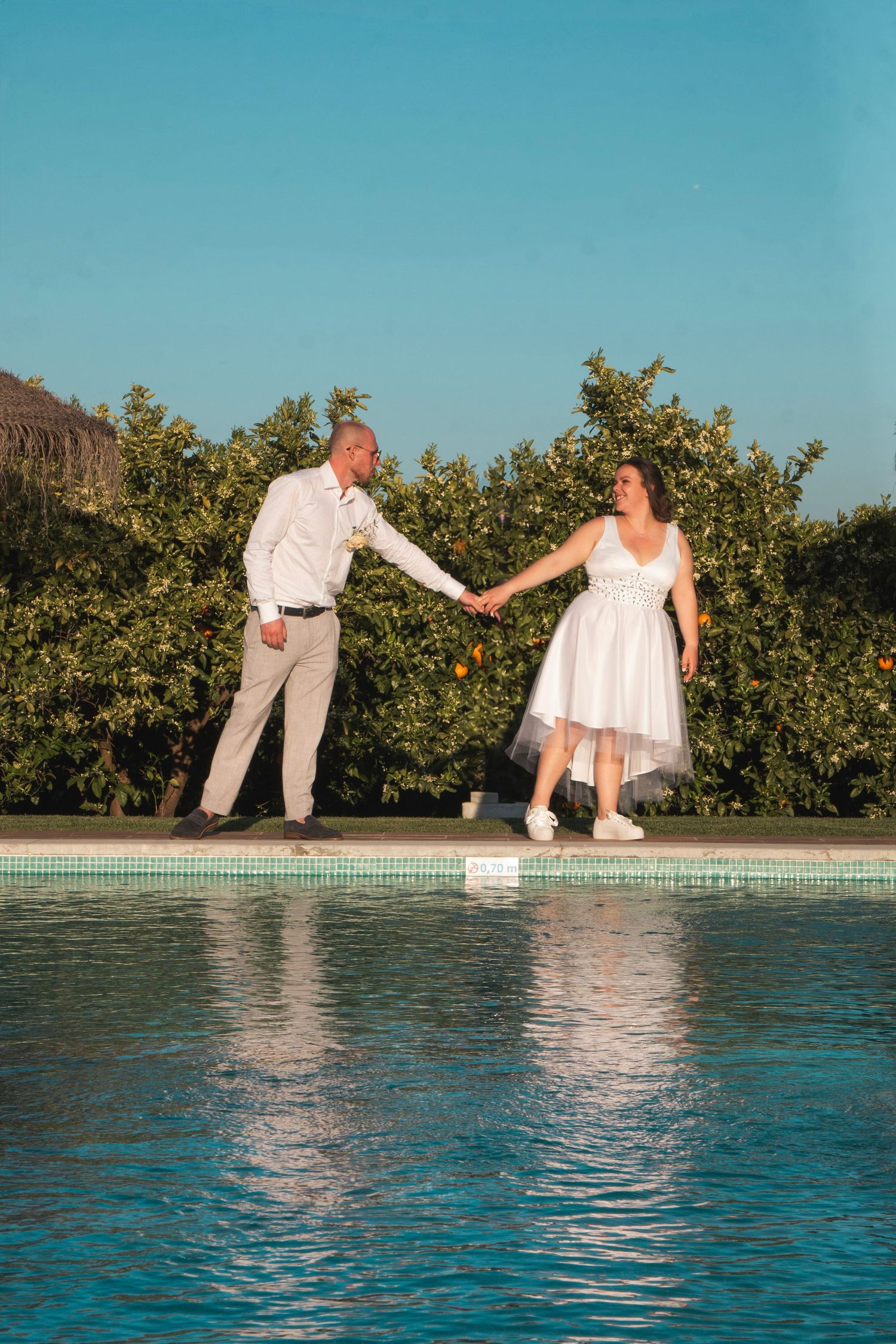 A couple in wedding attire holding hands by a swimming pool with mountains in the background.