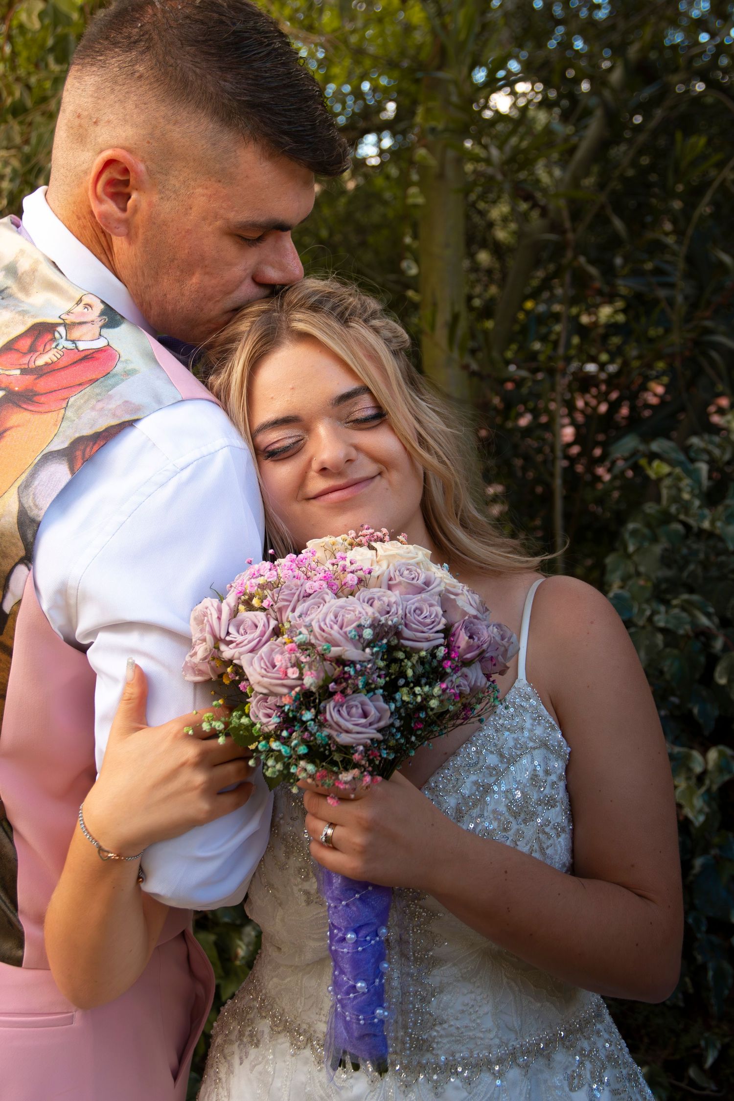 Bride and groom embrace tenderly outdoors, bride holding pink rose bouquet in lace dress as groom kisses her cheek.