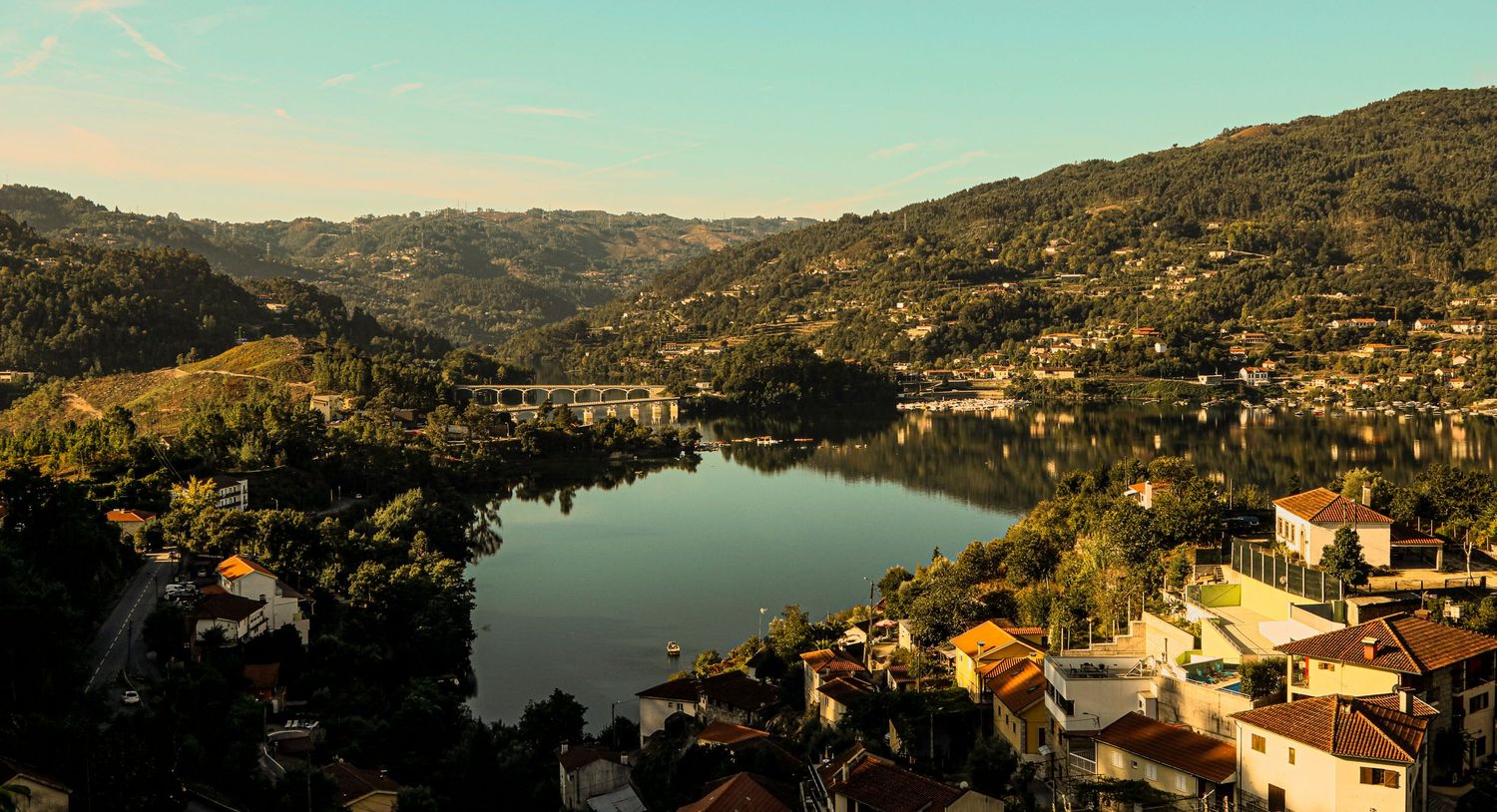 Scenic view of a mountain lake surrounded by hillside homes and lush terrain in golden afternoon light.