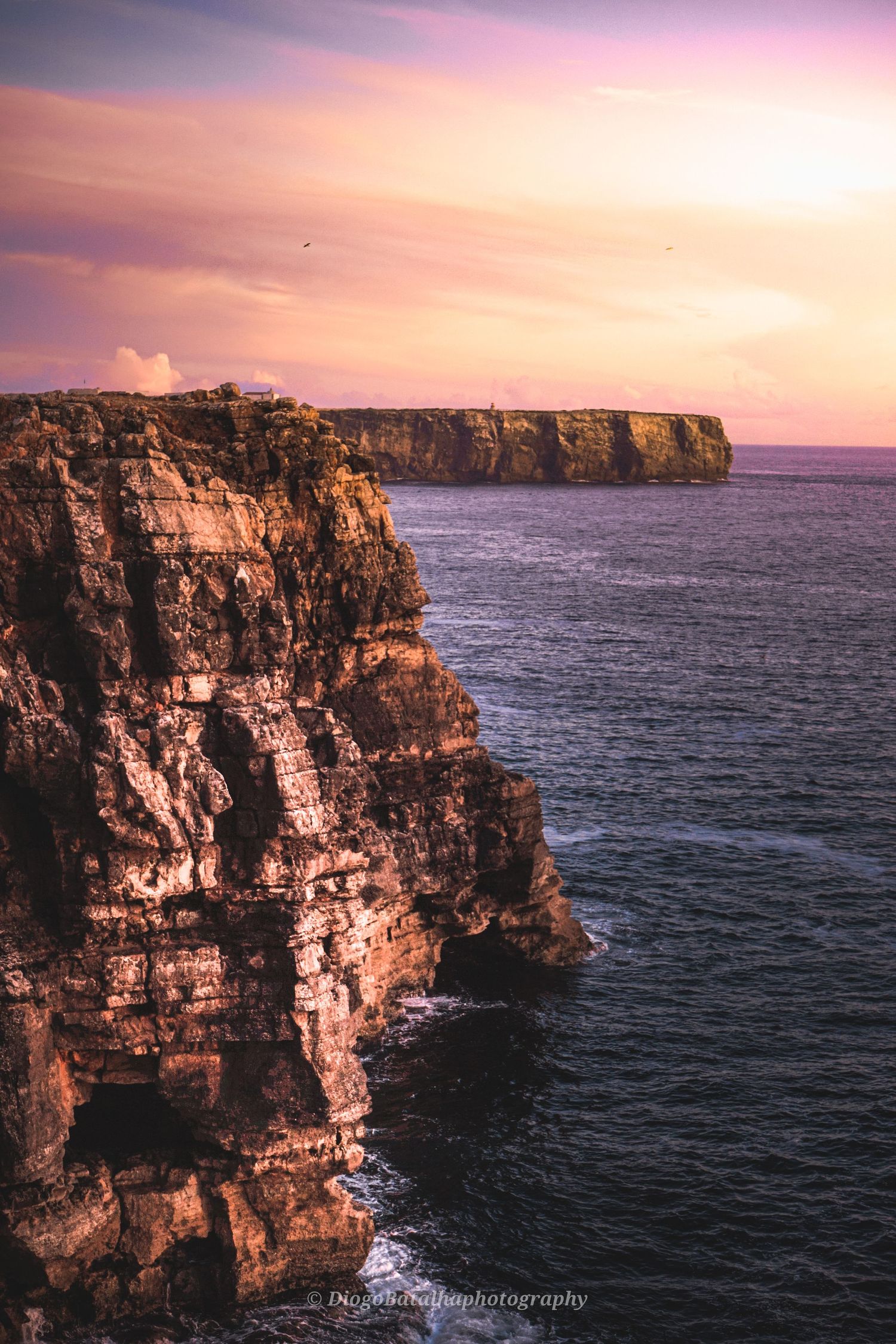 Dramatic coastal cliffs rise above the ocean at sunset with pink and purple sky illuminating the rocky landscape.