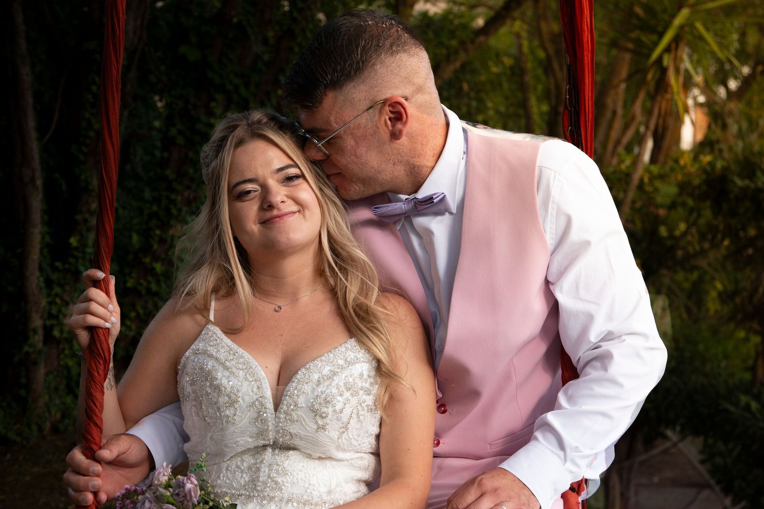 Bride in white lace gown sits on swing while groom in pink vest kisses her cheek at outdoor wedding.