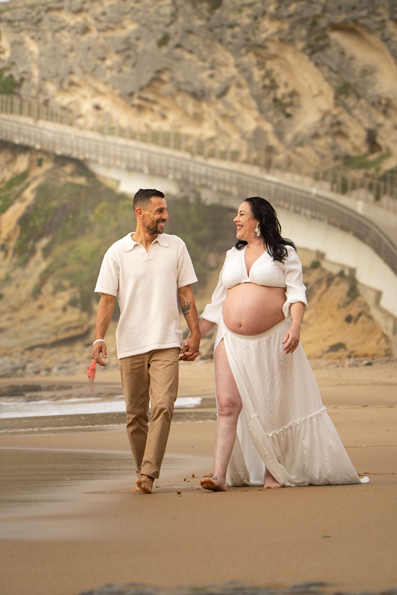 Pregnant couple walking hand in hand on sandy beach, woman in white dress showing baby bump, rocky cliffs in background.