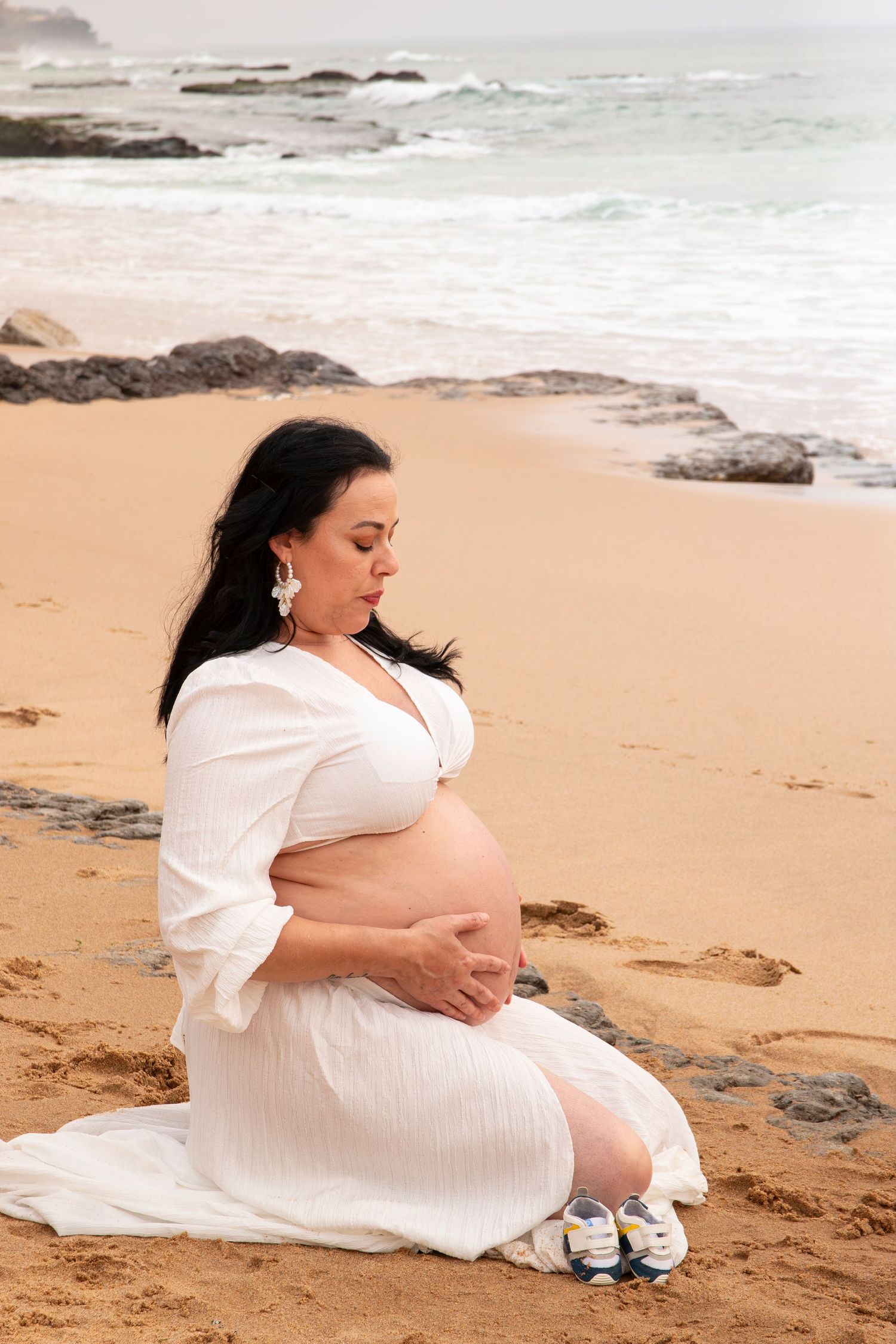 Pregnant woman in white dress sitting on sandy beach, cradling her belly near ocean waves with baby shoes beside her.