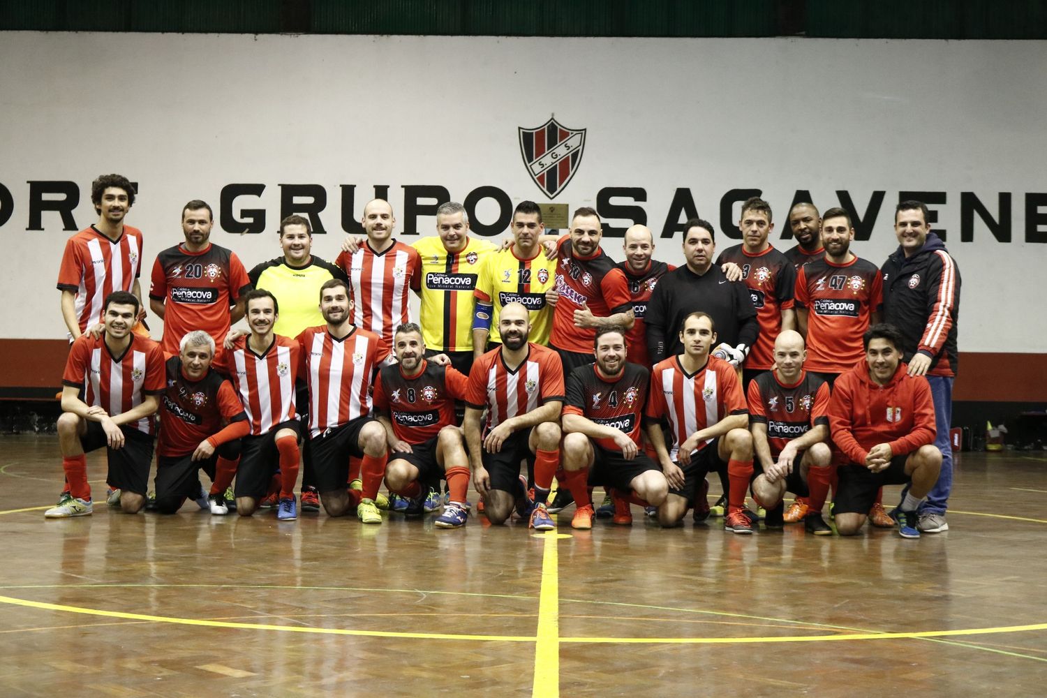 Indoor sports team poses for group photo wearing red and white striped jerseys on futsal court with banner behind them.