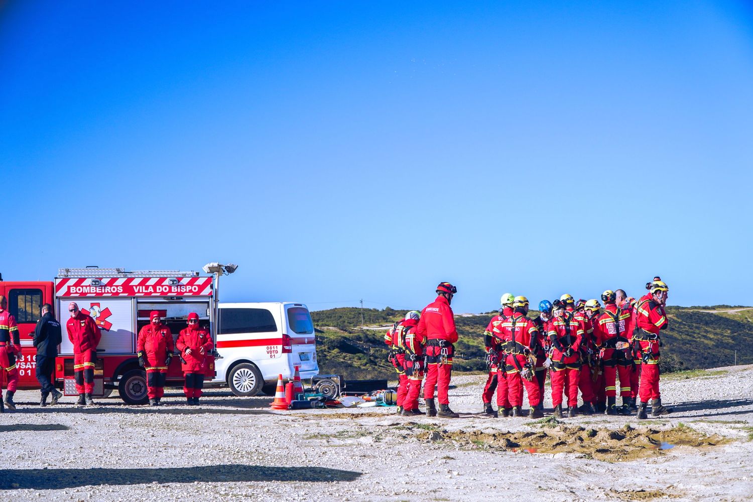 Group in red uniforms gathered on snowy terrain with vehicles in background.