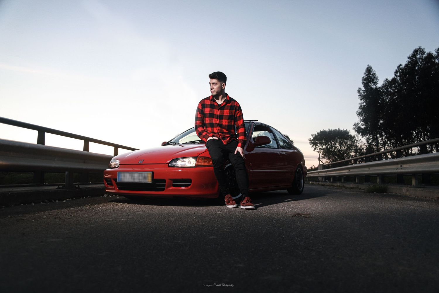 Red sporty tuned Honda Civic EG hatchback parked at sunset with person in plaid shirt leaning against hood.