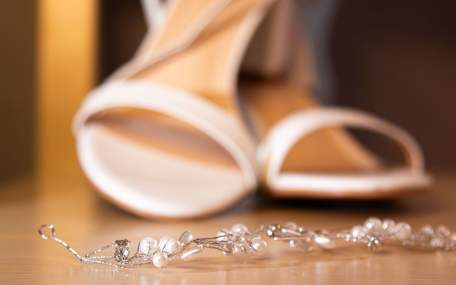Light colored bridal shoes with delicate pearl and crystal jewelry laid on wooden surface in soft focus.