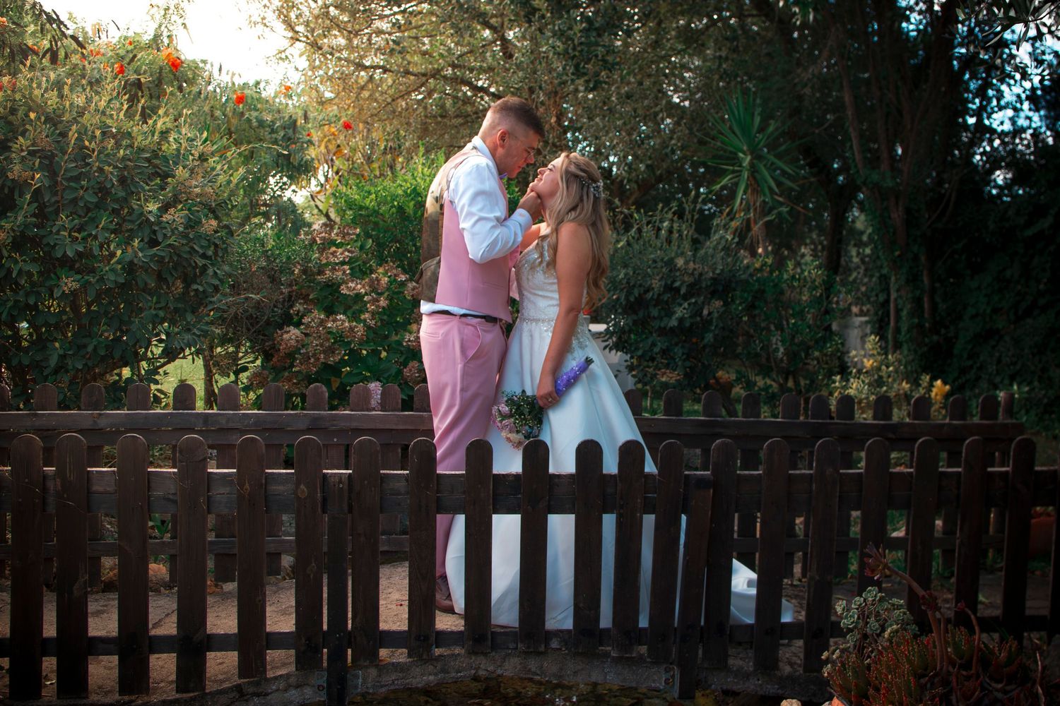 A couple in wedding attire shares a romantic kiss on a wooden bridge surrounded by lush green trees.