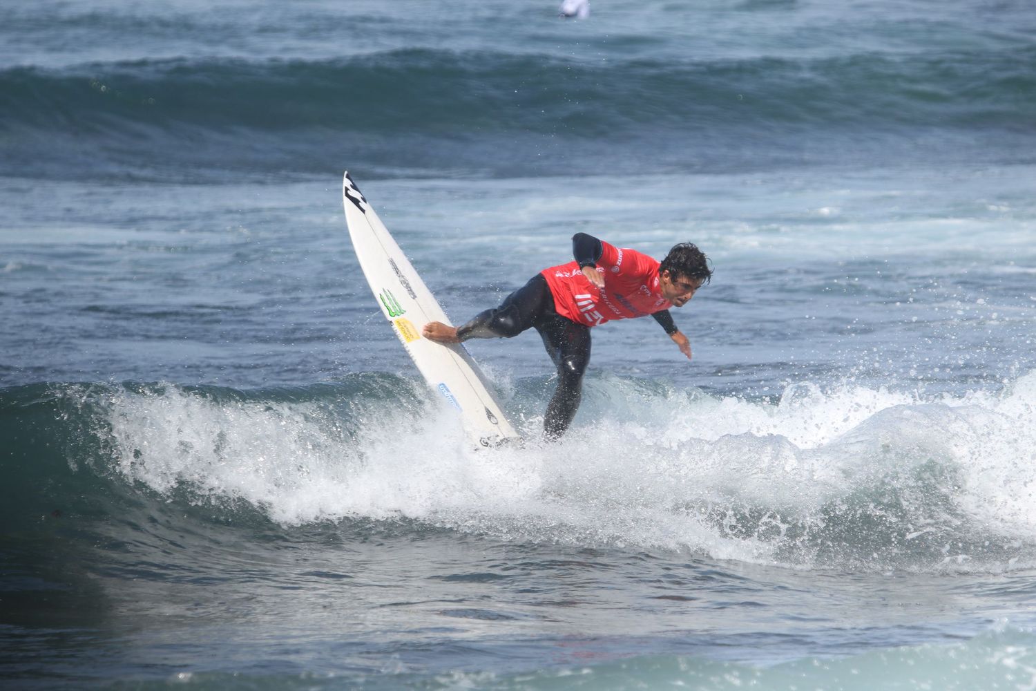 A surfer in a red wetsuit performs an aerial maneuver on a white surfboard over ocean waves.