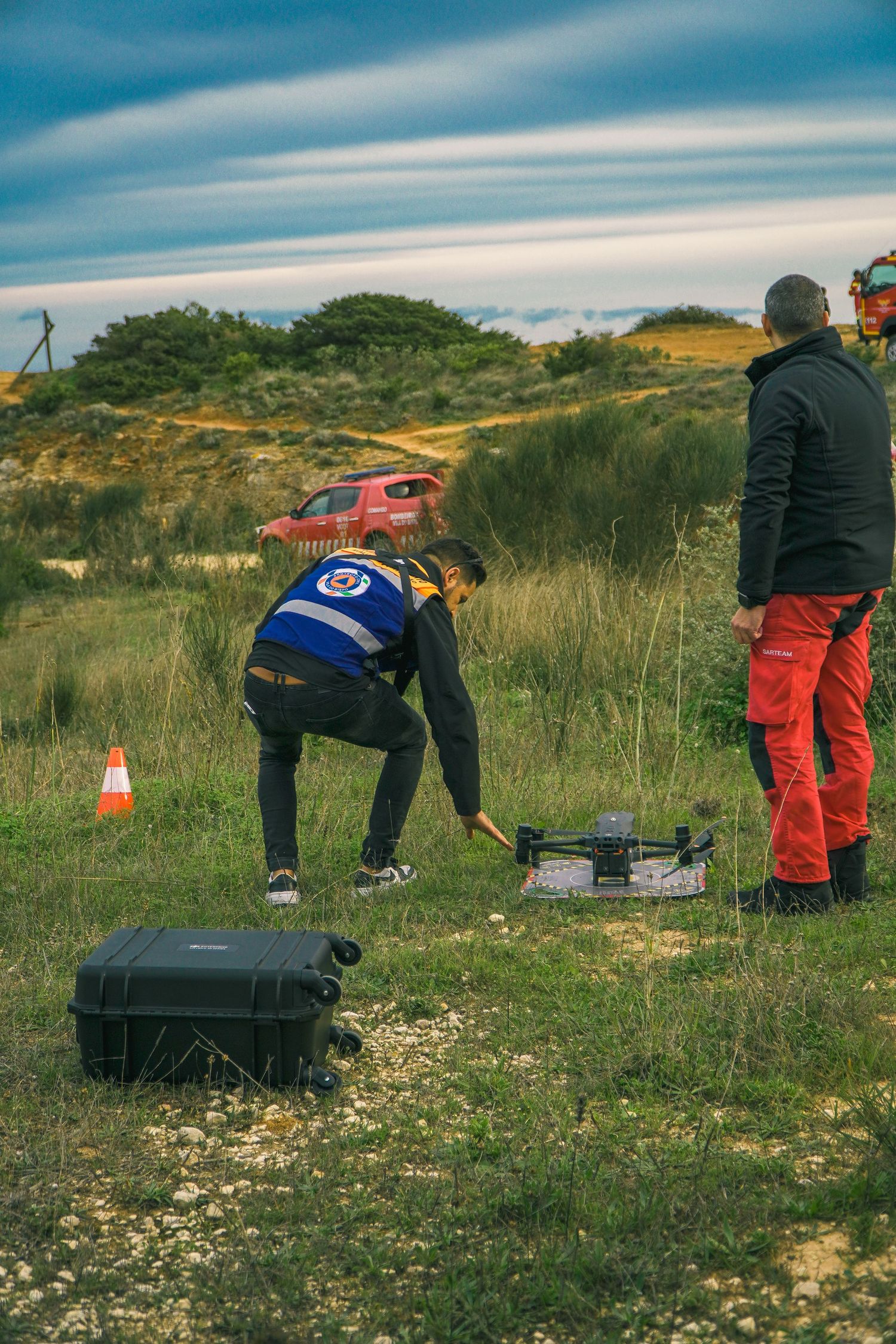 Three people in work attire crouch on grassy terrain while setting up equipment outdoors under a cloudy sky.