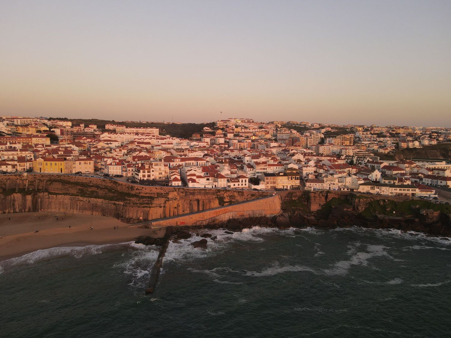 Aerial view of coastal town Ericeira in Portugal with historic buildings and cliffs along the Atlantic Ocean at sunset.