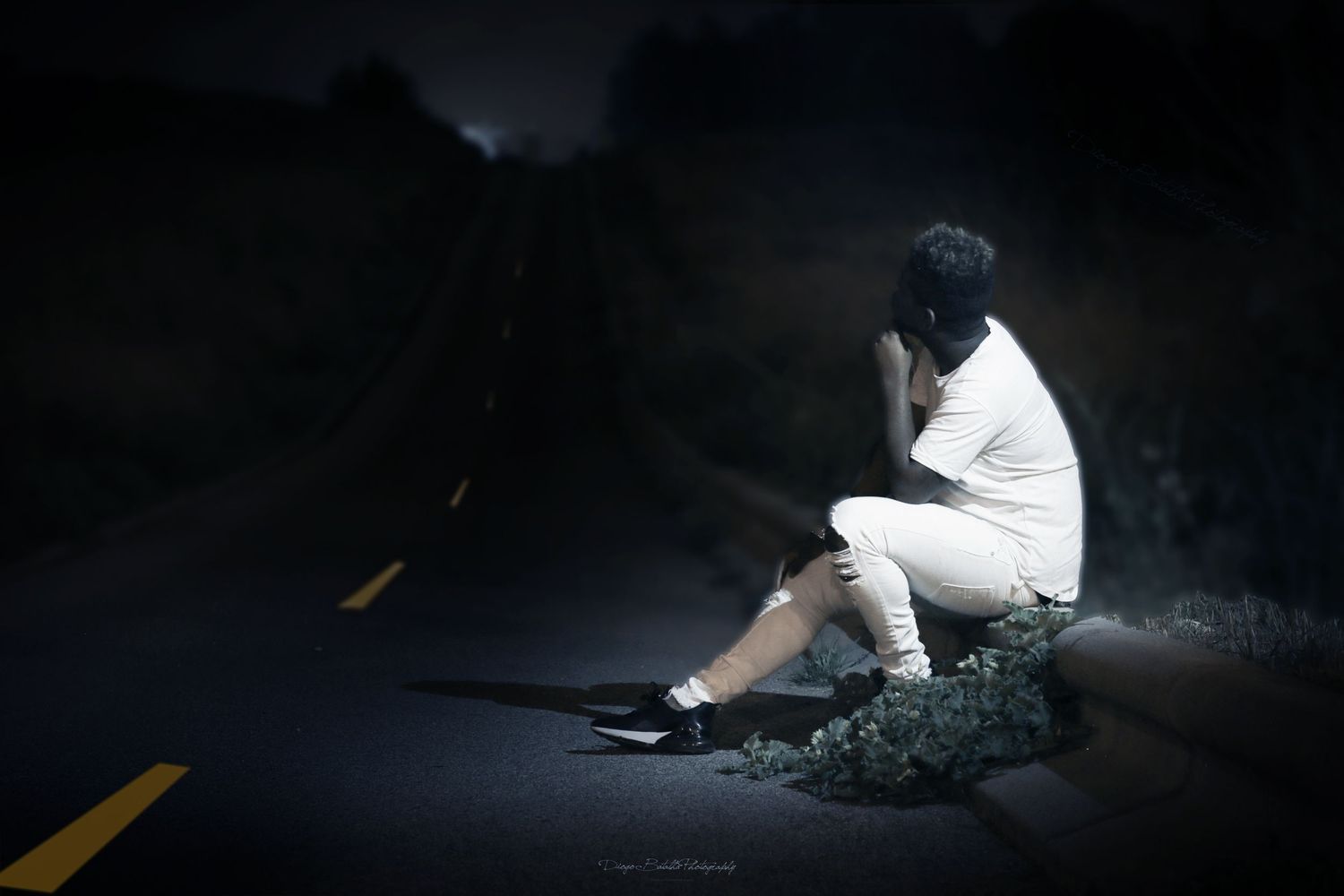 A person in white clothing sits alone on a rock at night on a dark road with dramatic lighting.