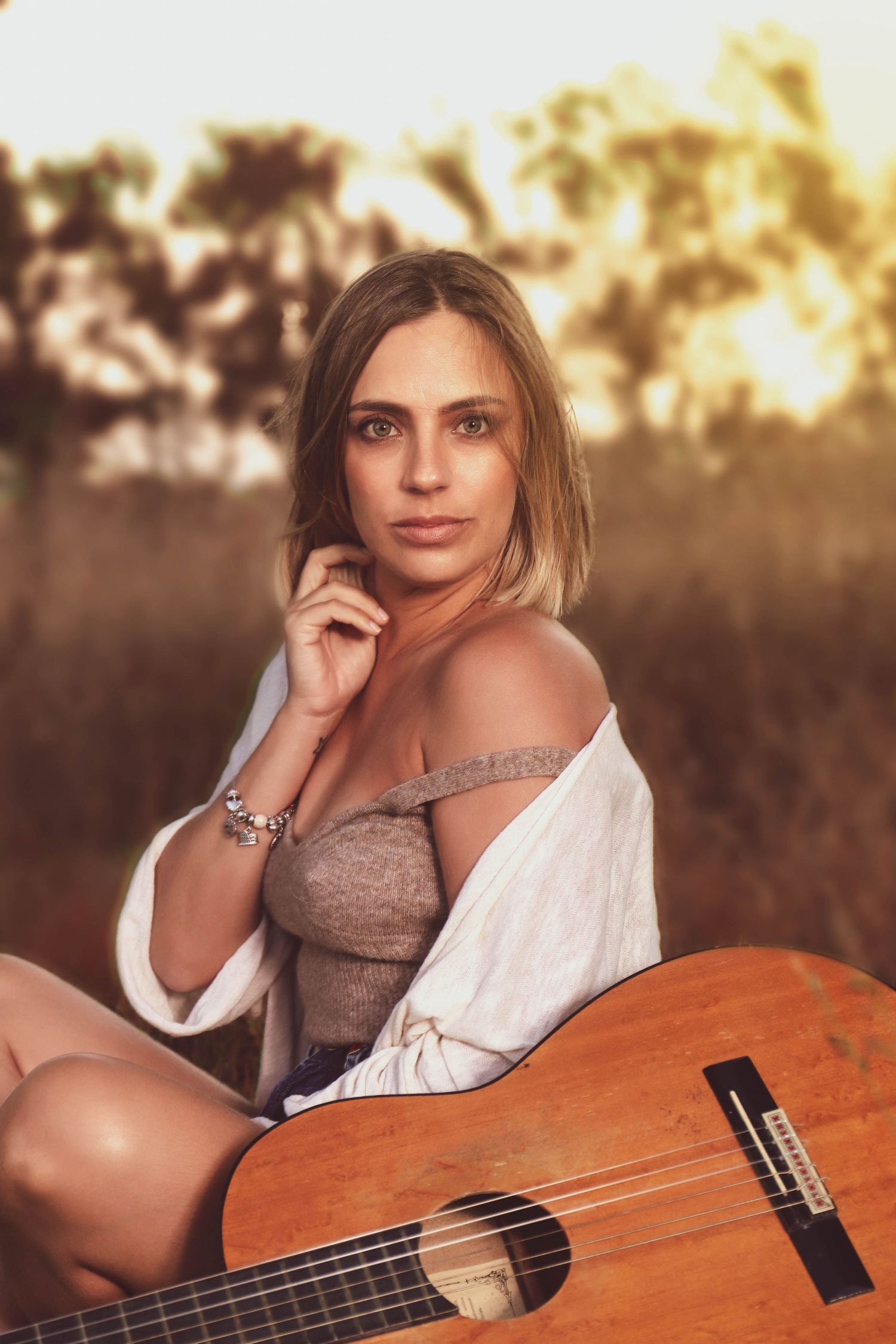A seated musician poses outdoors at sunset with an acoustic guitar, wearing a beige corset top and white cardigan.
