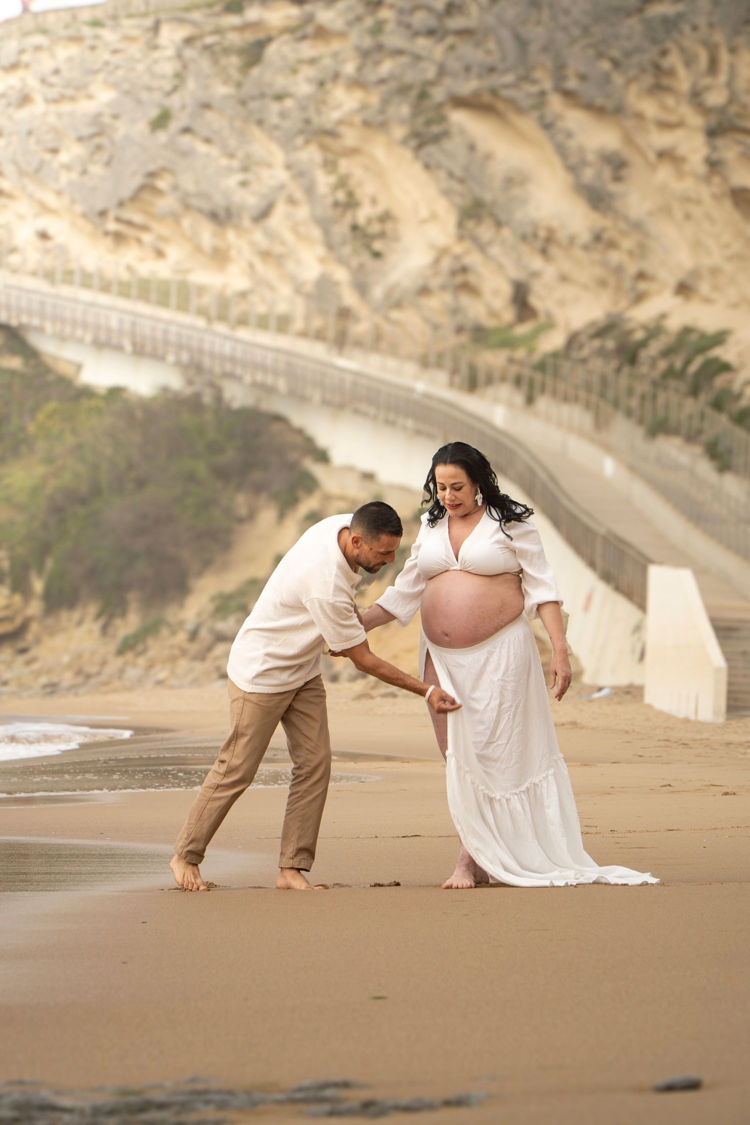 Pregnant woman in white dress with partner on sandy beach, rocky cliffs and bridge in background.