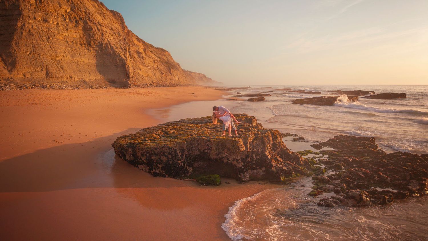 Wide panoramic shot of a rugged beach coastline with orange cliffs and crashing waves at sunset.