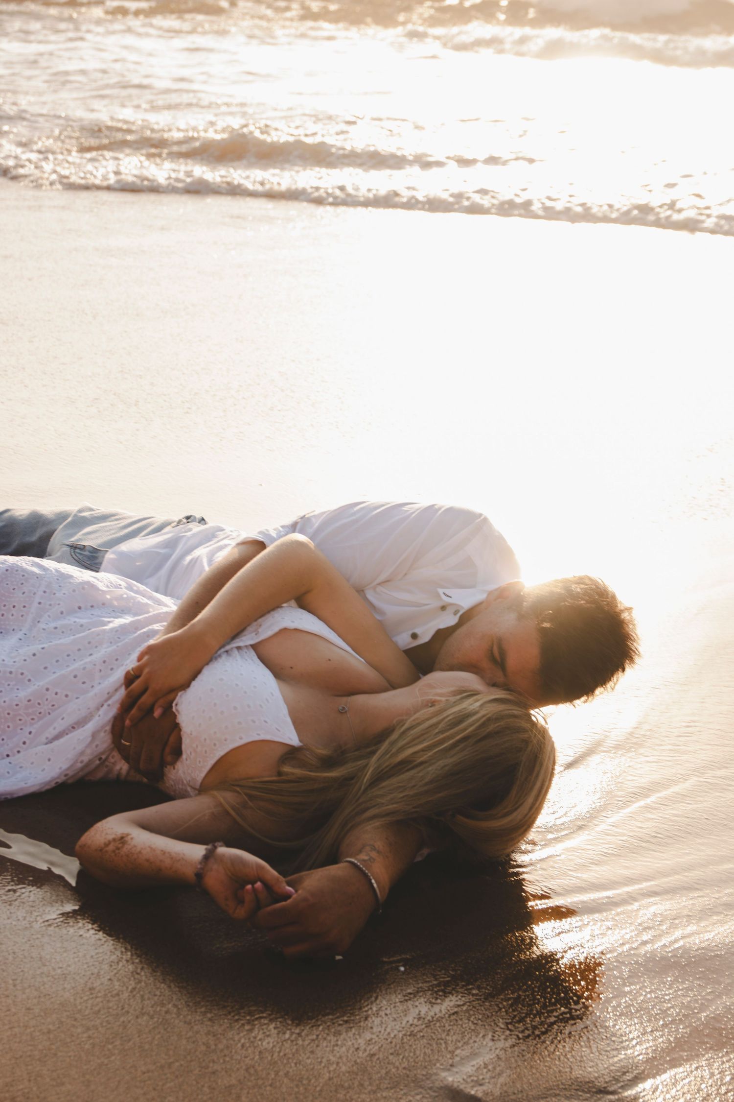 Ethereal figure in white dress lying on wet beach sand during golden hour at sunset.