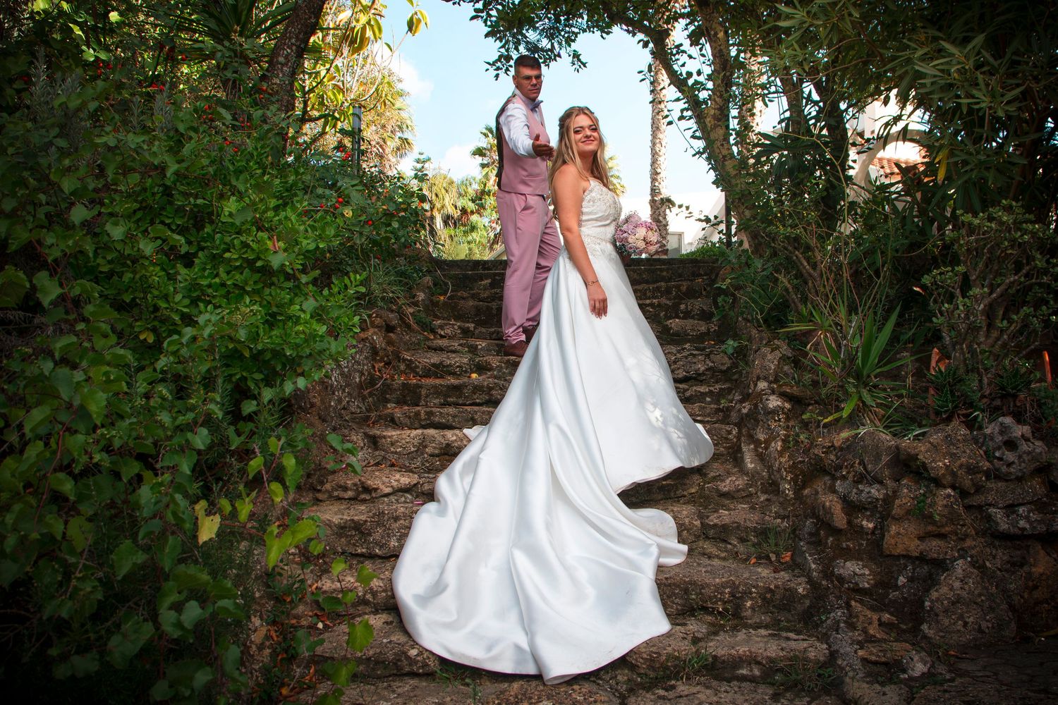 Bride in white gown with long train poses on stone steps with groom in pink suit amid lush greenery.