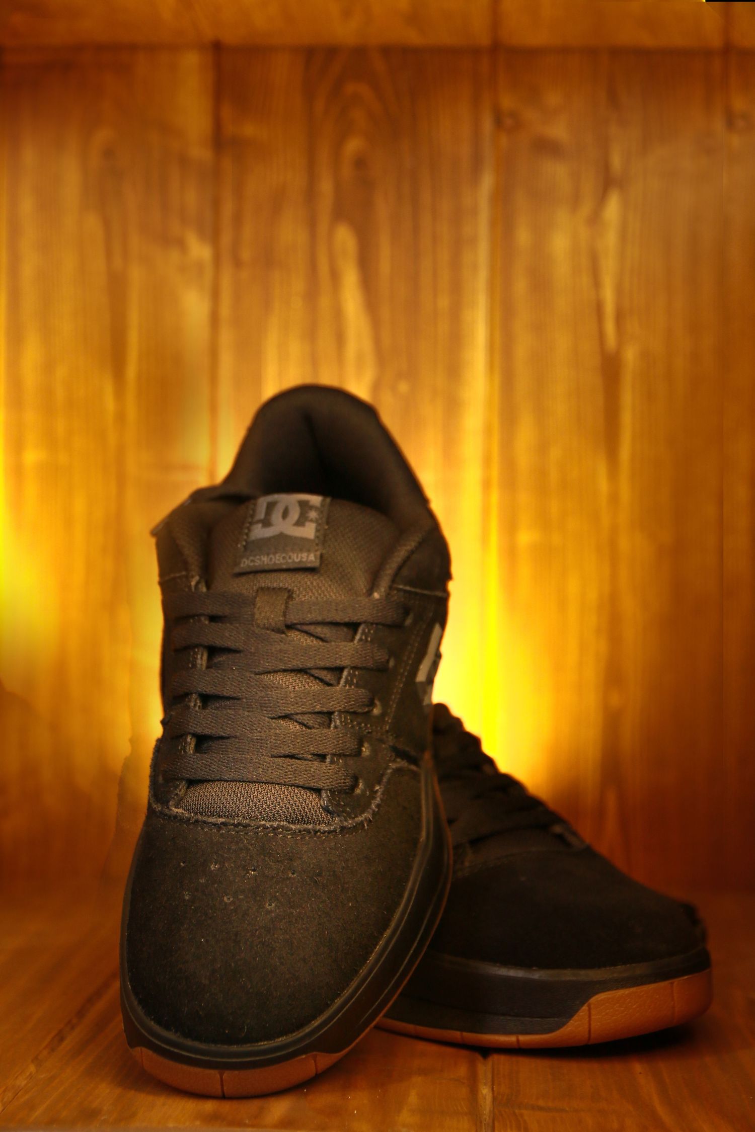 Black DC skate shoes with gum sole photographed against warm wooden background with dramatic lighting.