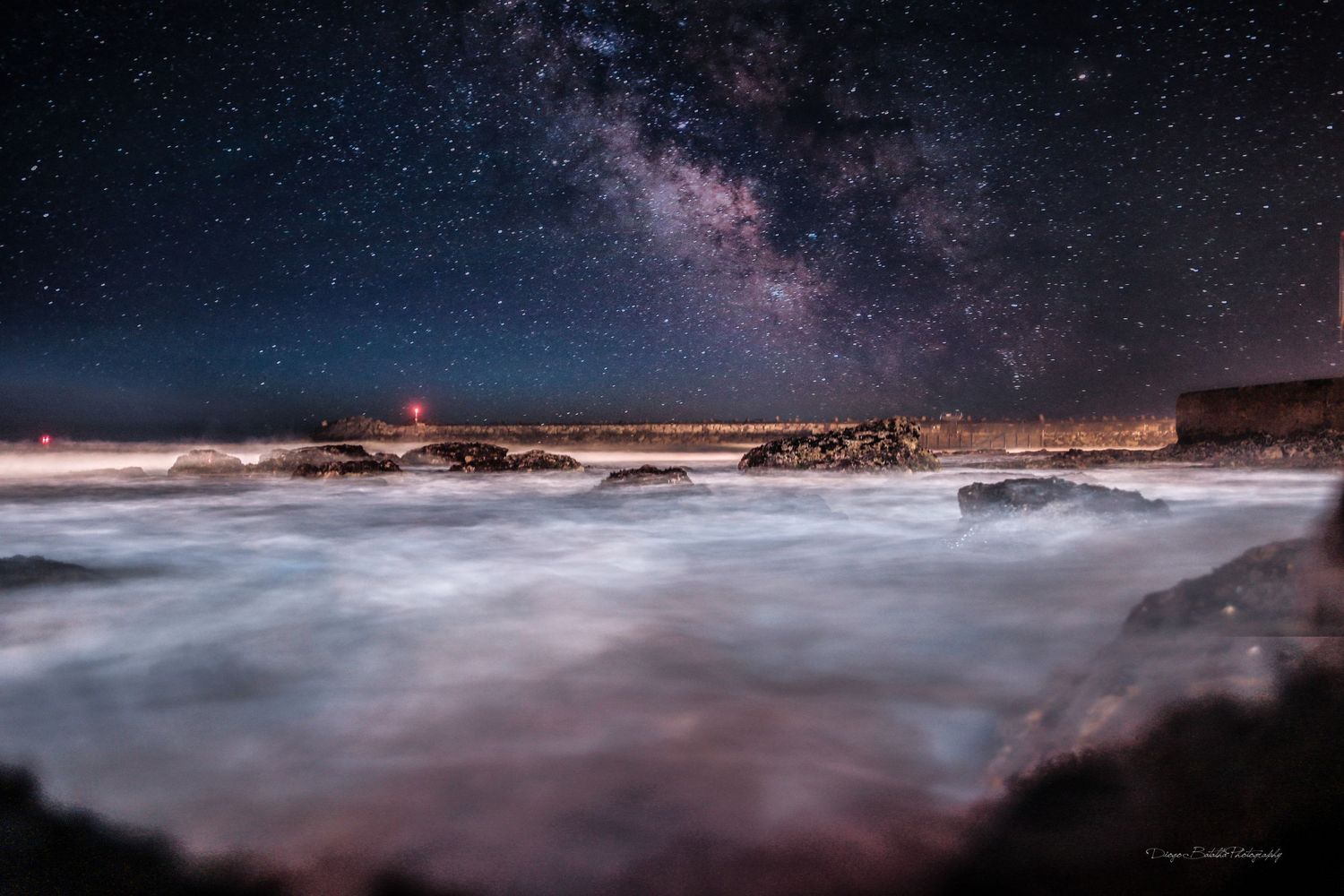 Misty ocean waves crash against rocky coast under starlit Milky Way galaxy in night sky.