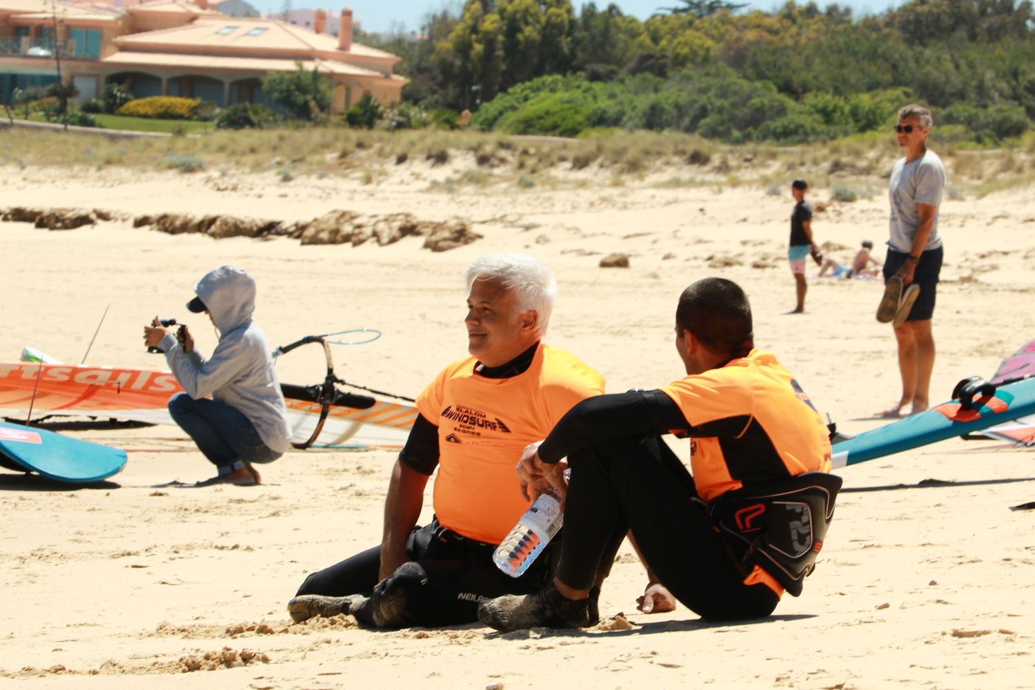Students sitting on the beach during a windsurfing lesson with colorful surfboards nearby.