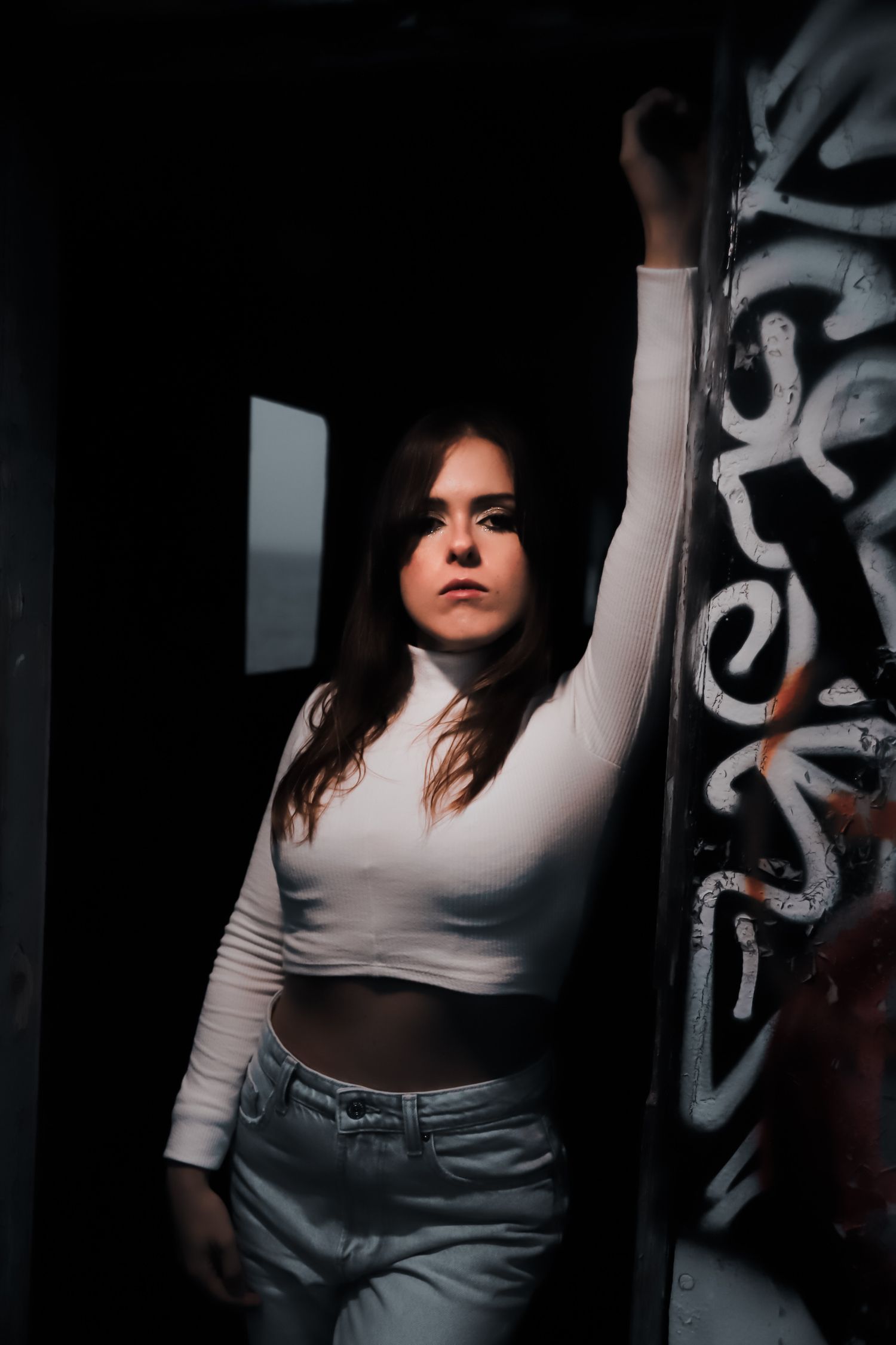 Dramatic portrait shot in dark room showing person in white crop top and jeans posing against wall with graffiti.