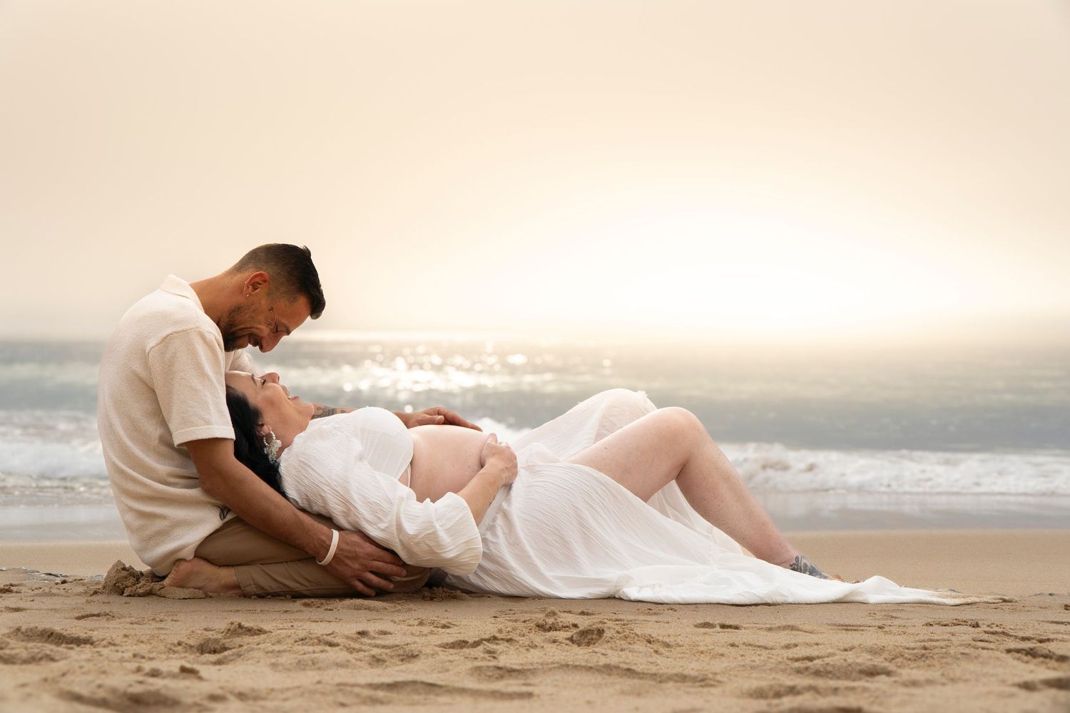 Pregnant couple sharing a tender moment on a sandy beach at golden sunset, woman in white dress lying back.