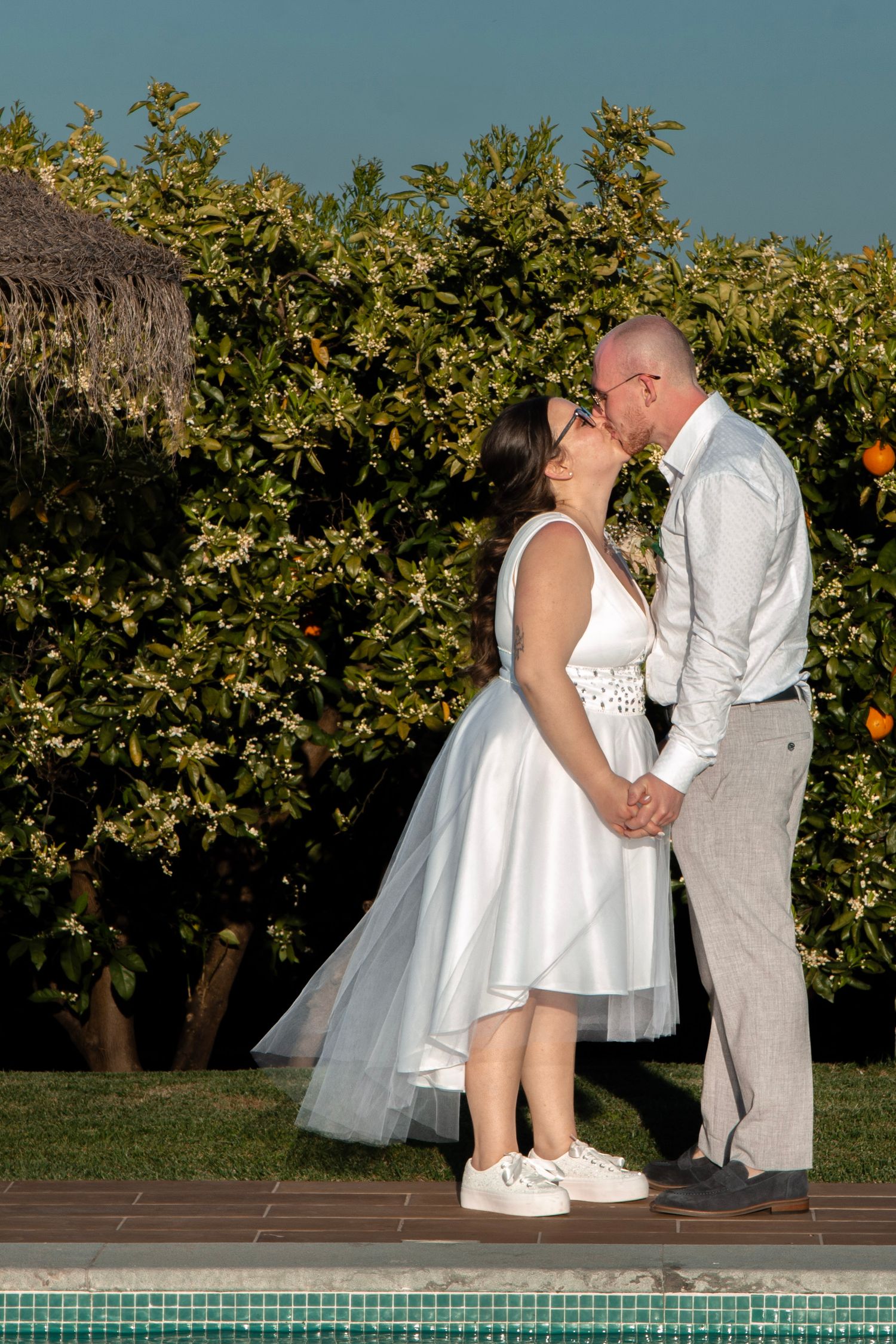 Newlyweds share a kiss by pool with citrus grove in background.