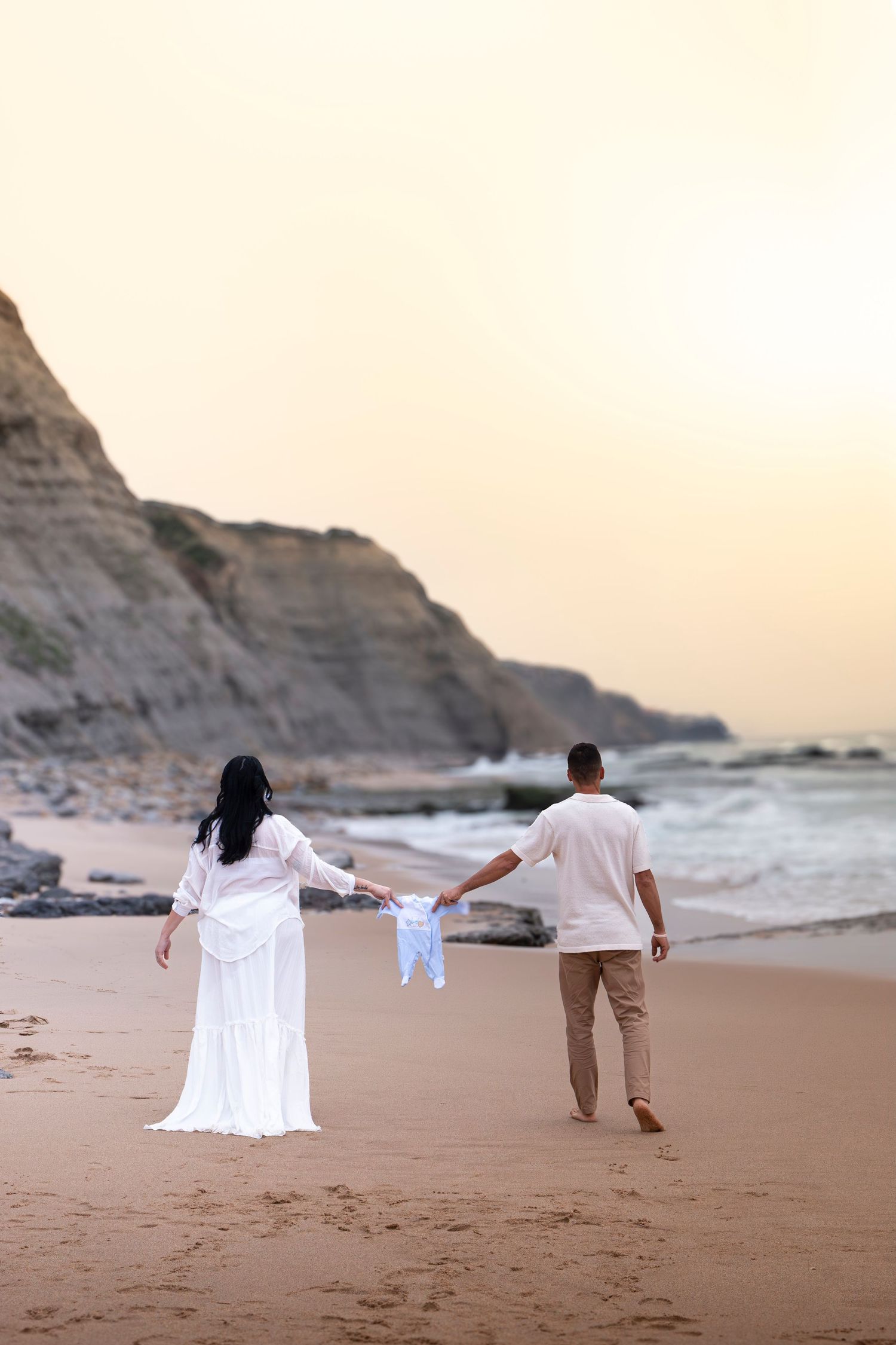 Couple in white walking on sandy beach at sunset, holding baby clothes between them with rocky cliffs in background.