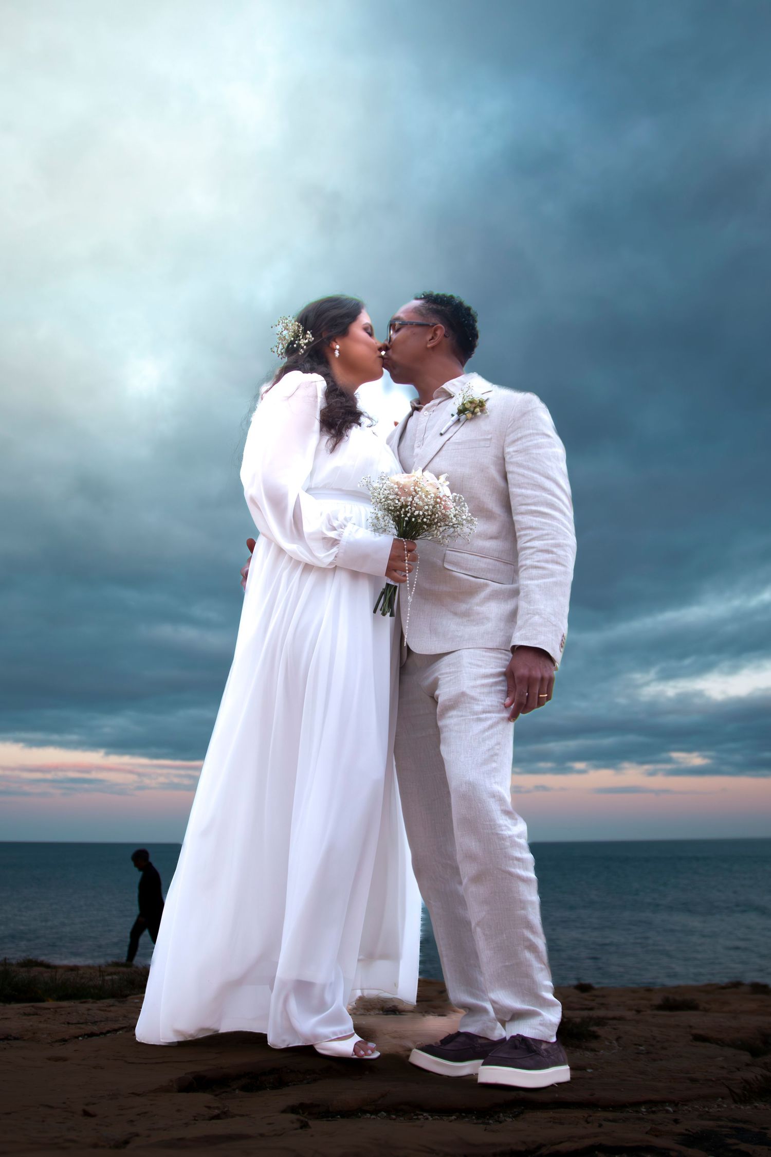 Wedding couple in white embraces on a dramatic beach against stormy skies during sunset.