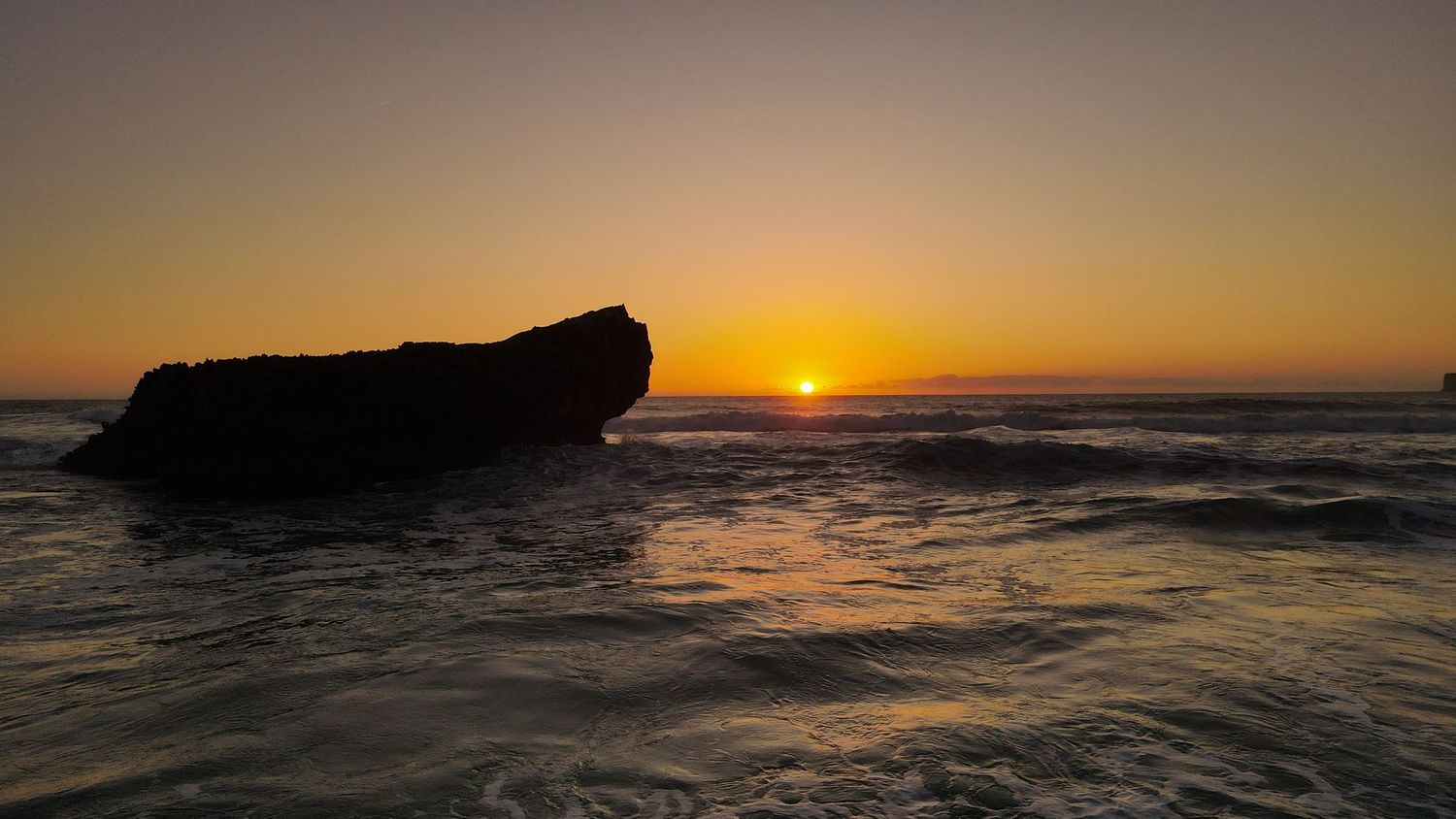 A large rock formation silhouetted against a vibrant orange sunset over the ocean waves.