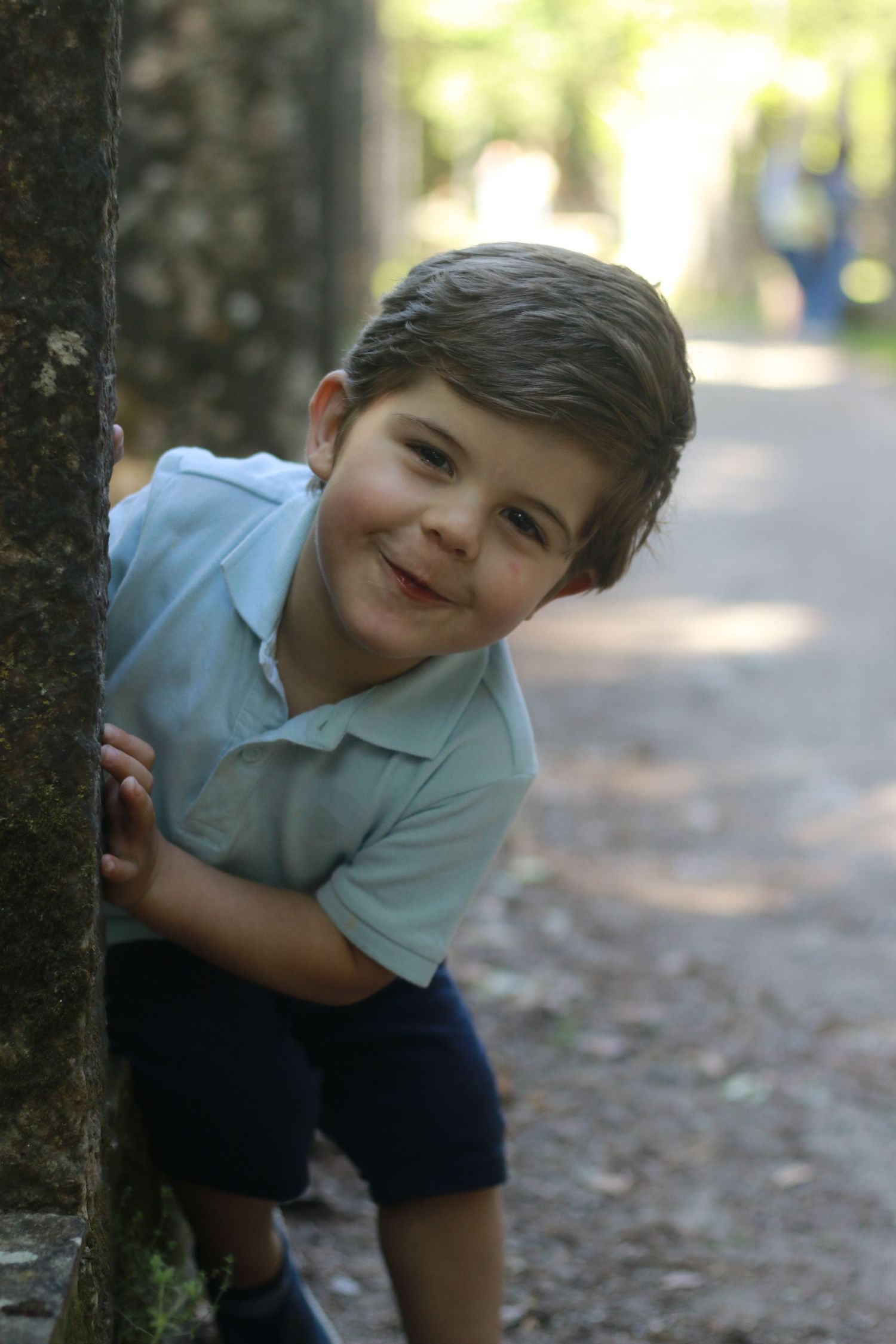 Young child in light blue polo peeks playfully from behind tree trunk on forest path.