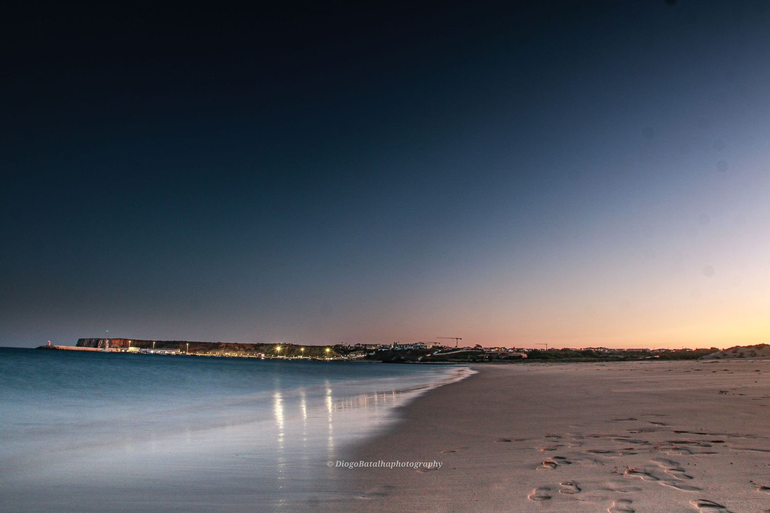 Beach shoreline at dusk with distant lights reflecting on calm waters.