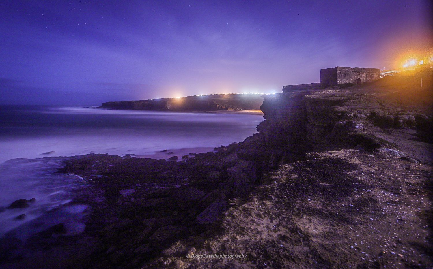 Dramatic purple twilight over a rocky coastal cliff with distant lights twinkling along the shoreline and buildings silhouetted.