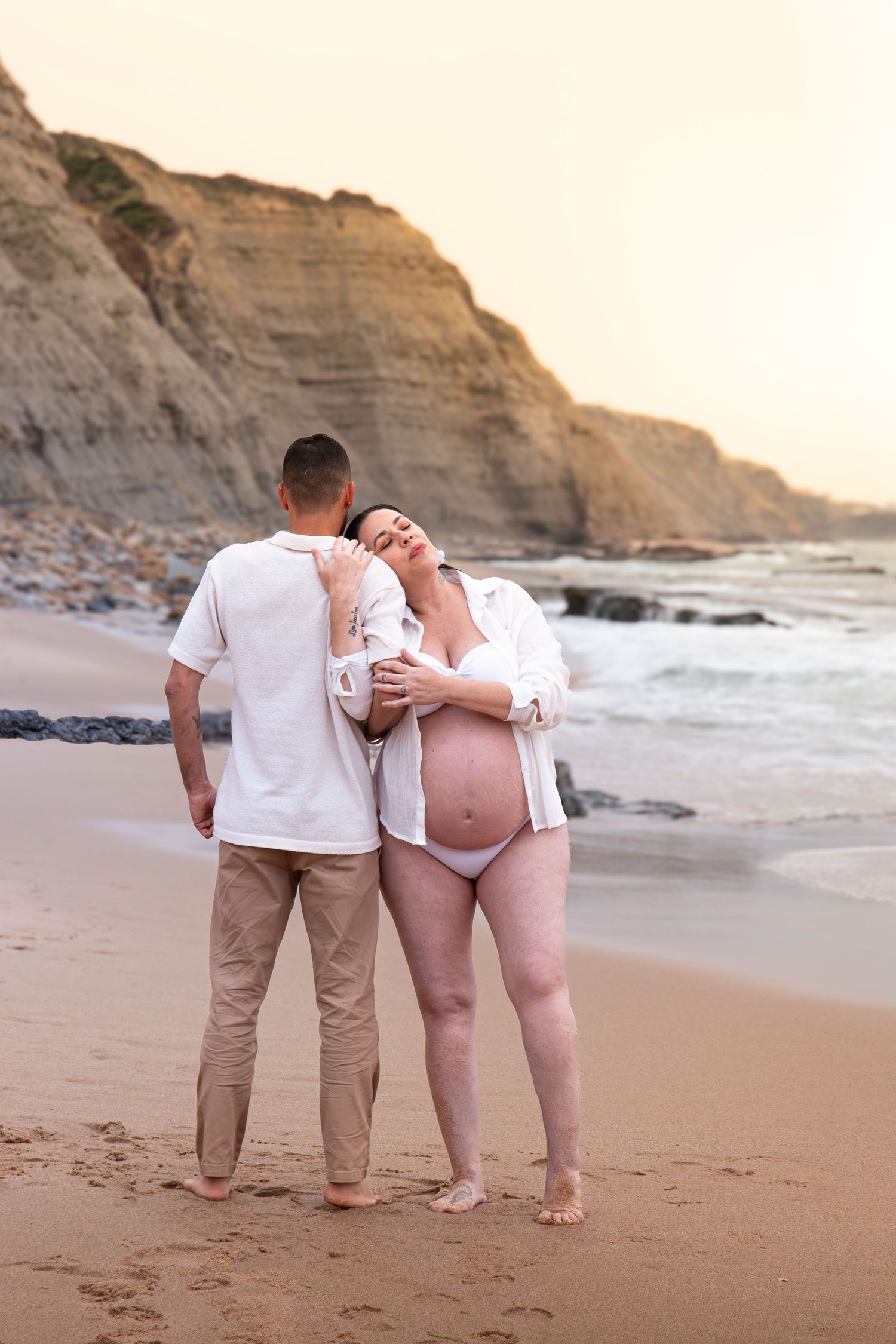 Pregnant couple embracing on a sandy beach at sunset with rocky cliffs in the background.