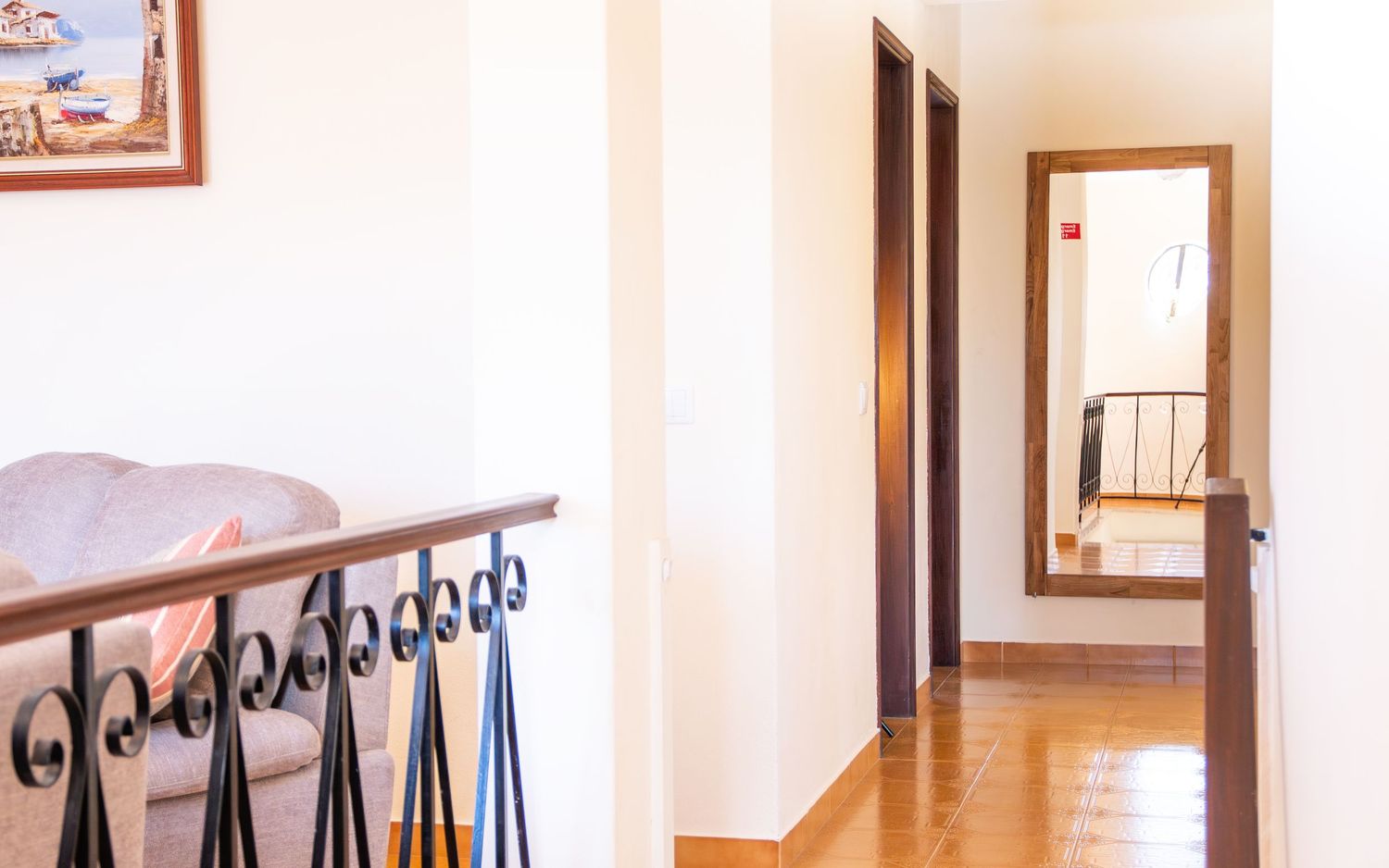 Interior hallway with hardwood floors, white walls, and decorative iron railing leading to doorway with mirror.