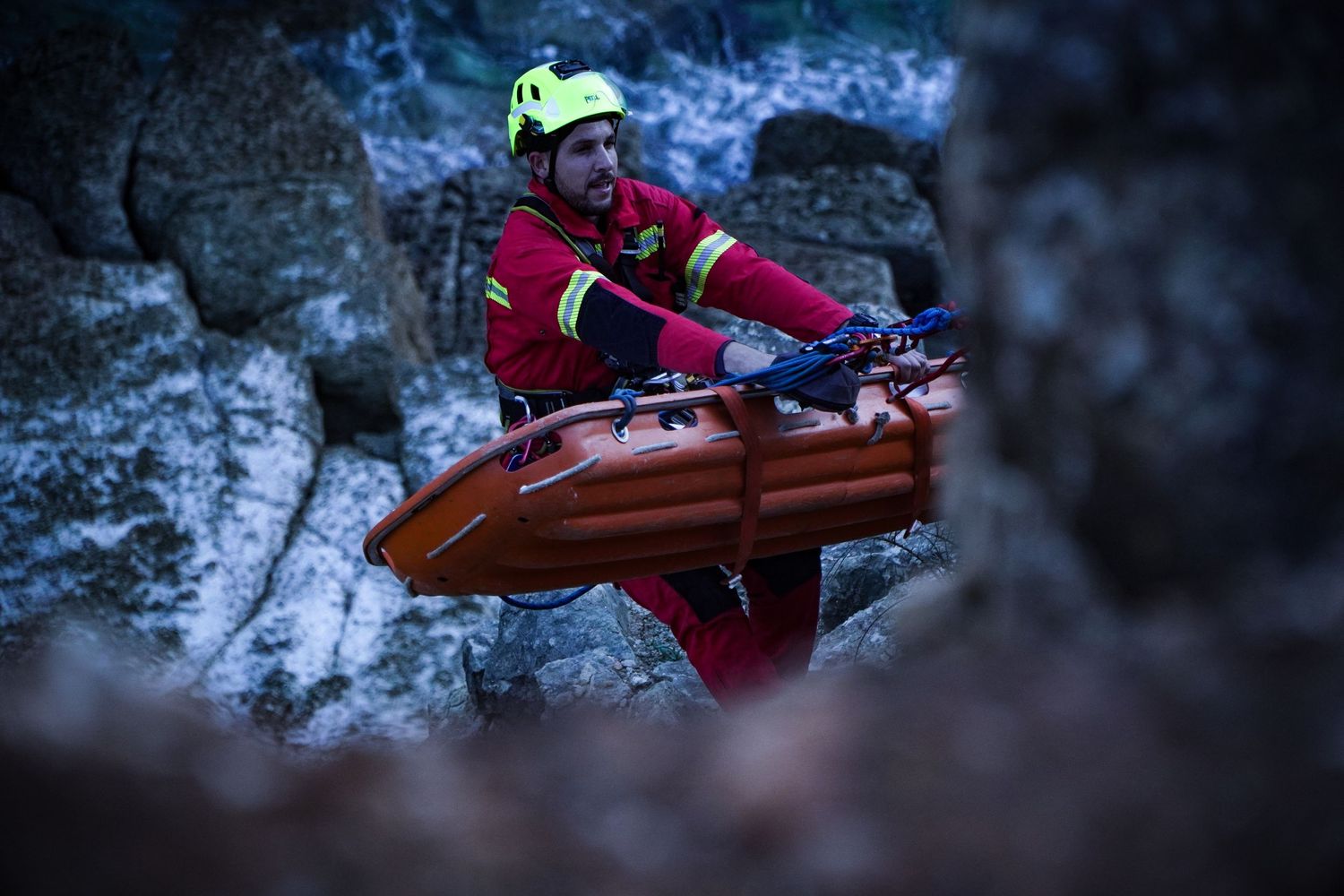 A rescue worker in red gear maneuvers a stretcher through a dark rocky terrain at night.