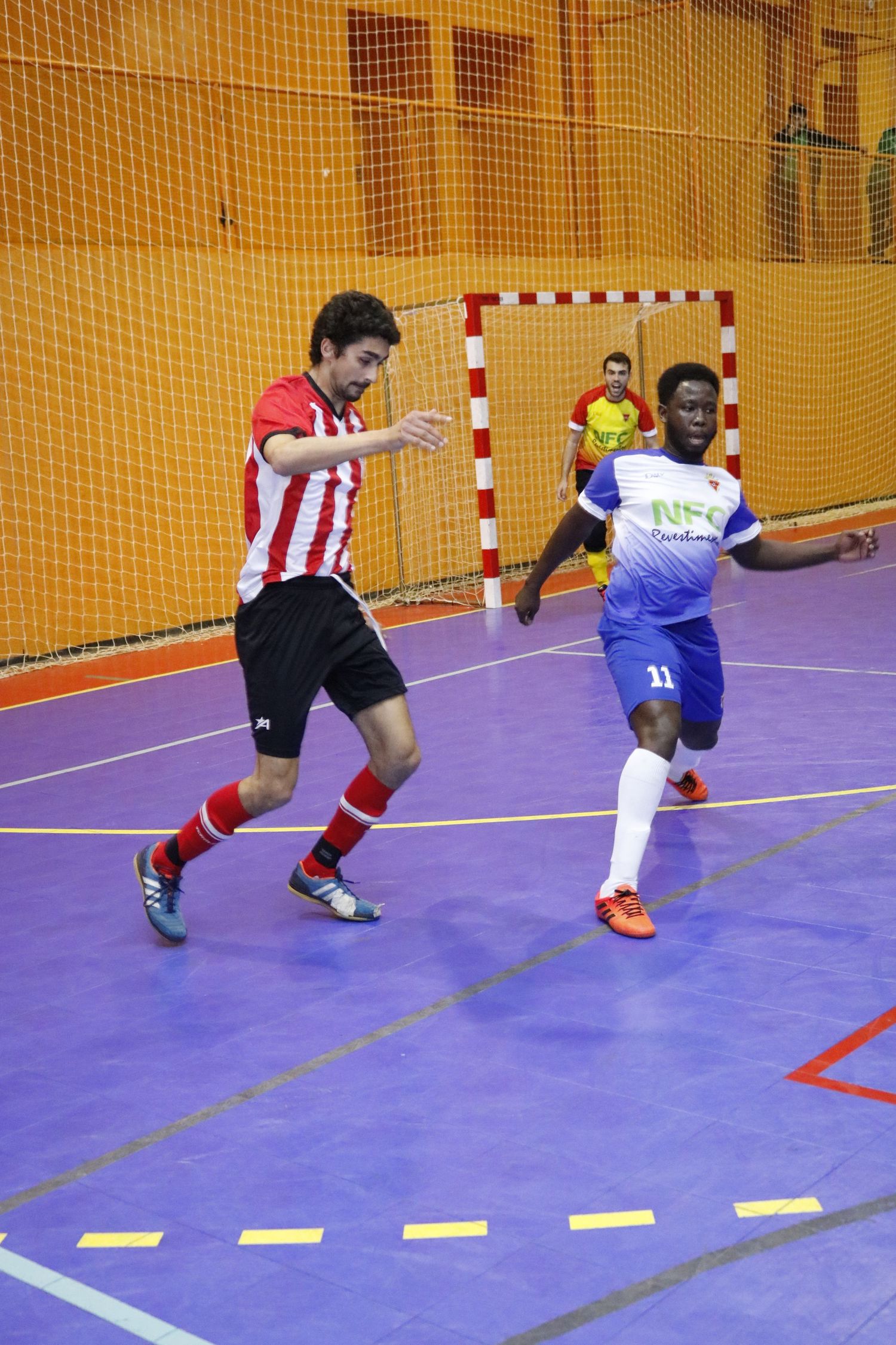 Indoor futsal match between players in red-white striped and blue-white uniforms on purple court with yellow netting.