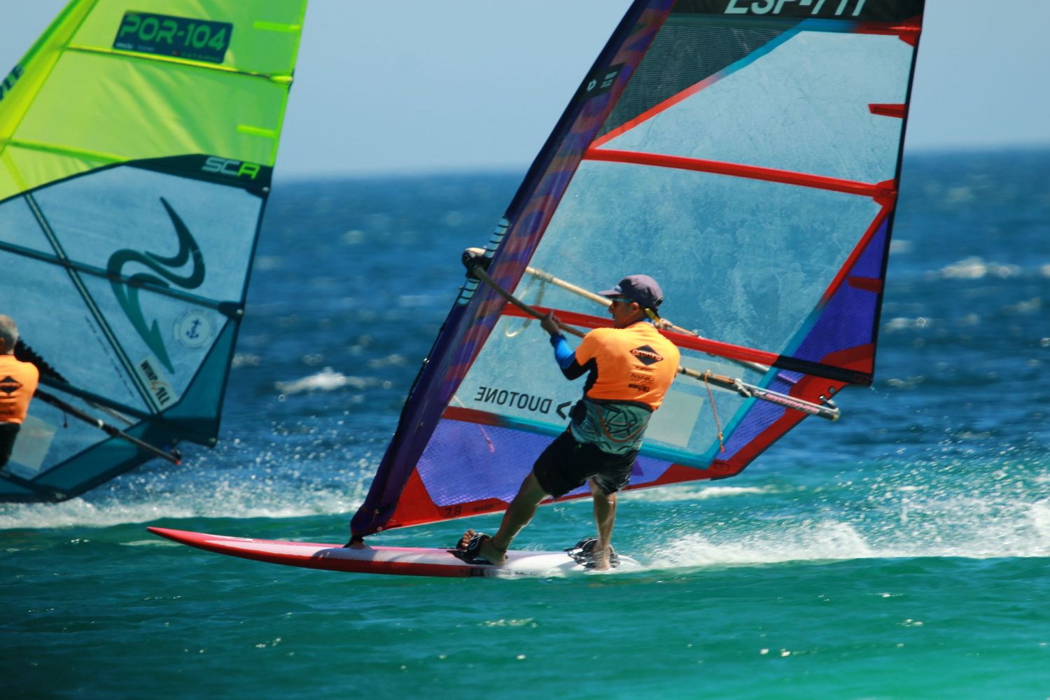 Windsurfer in orange riding across blue waters with colorful sails in action.