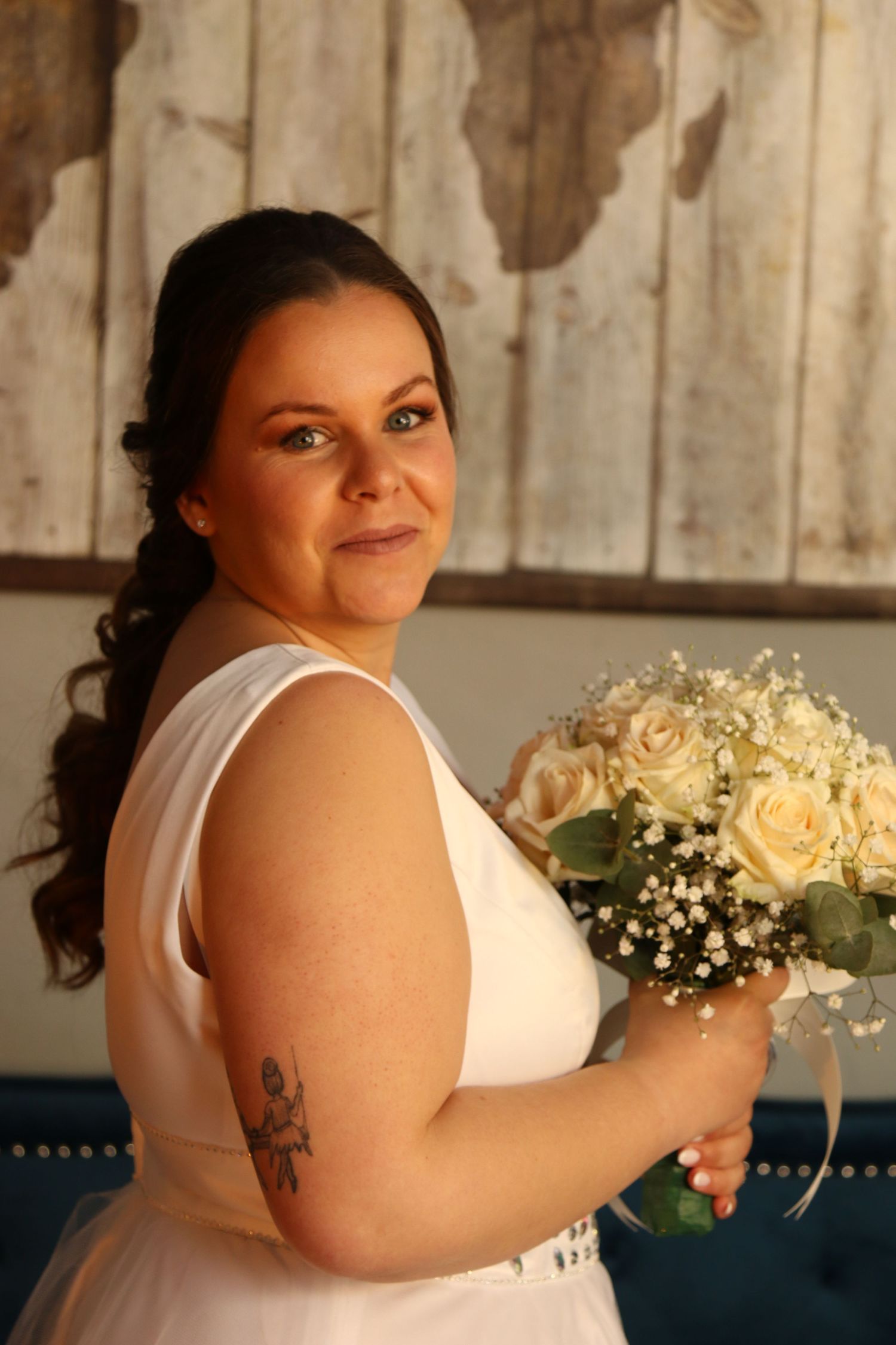 A bride in a white sleeveless dress holds a cream rose bouquet in front of a rustic wood wall backdrop.