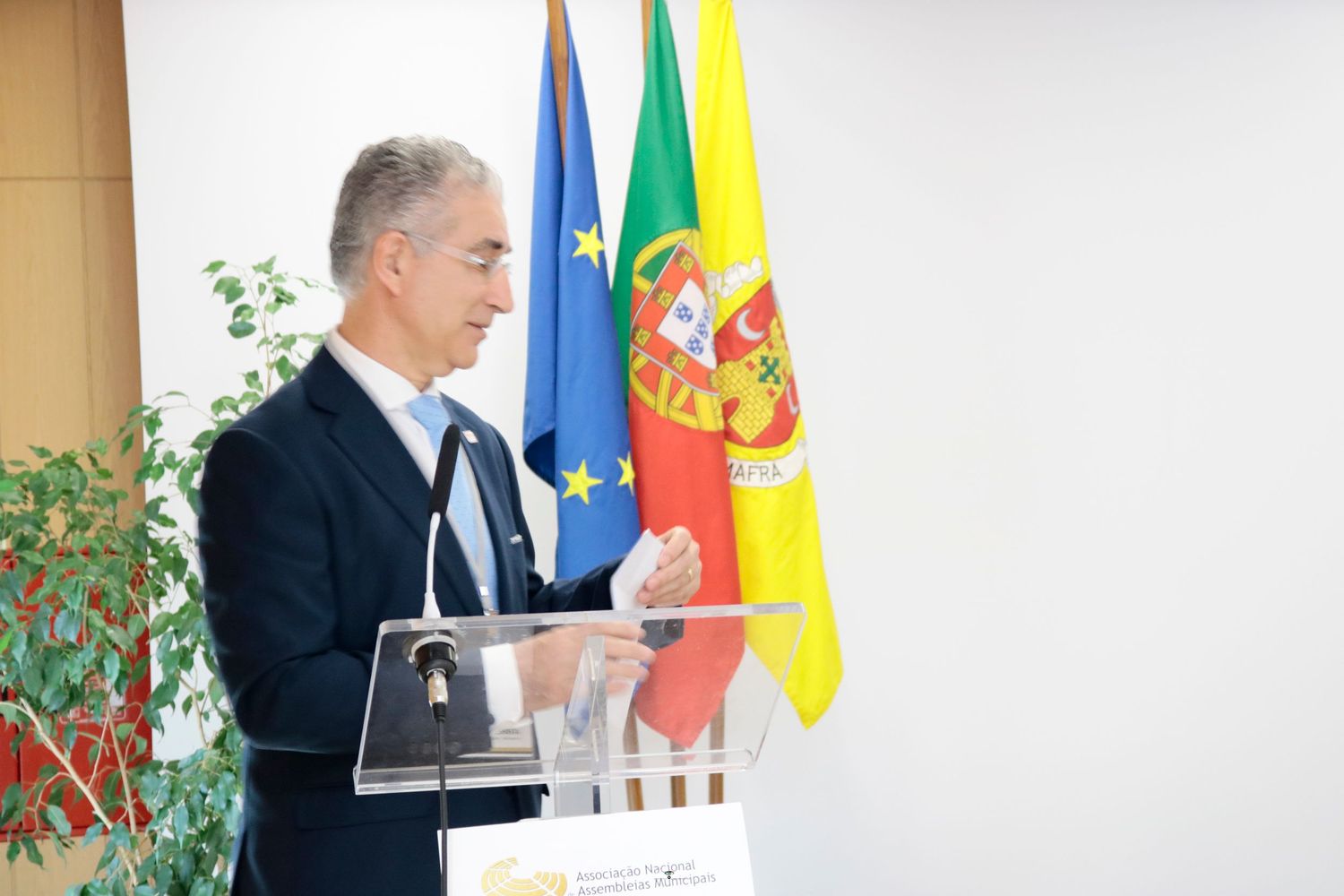 Speaker stands at podium speaking during diplomatic meeting with Portuguese and European Union flags visible in background.