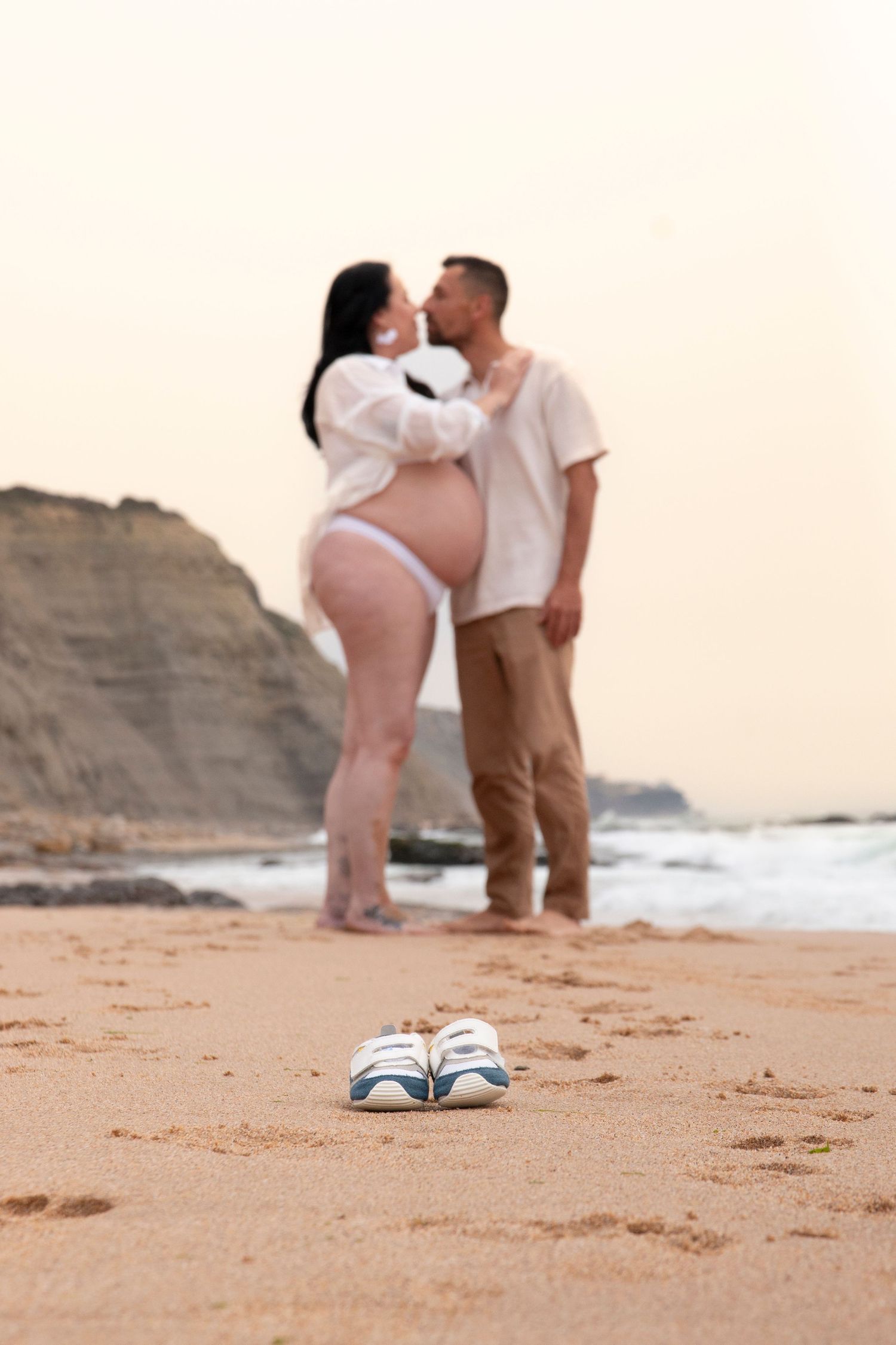 Pregnant couple kissing on beach with baby shoes in foreground, romantic maternity photo at sunset.