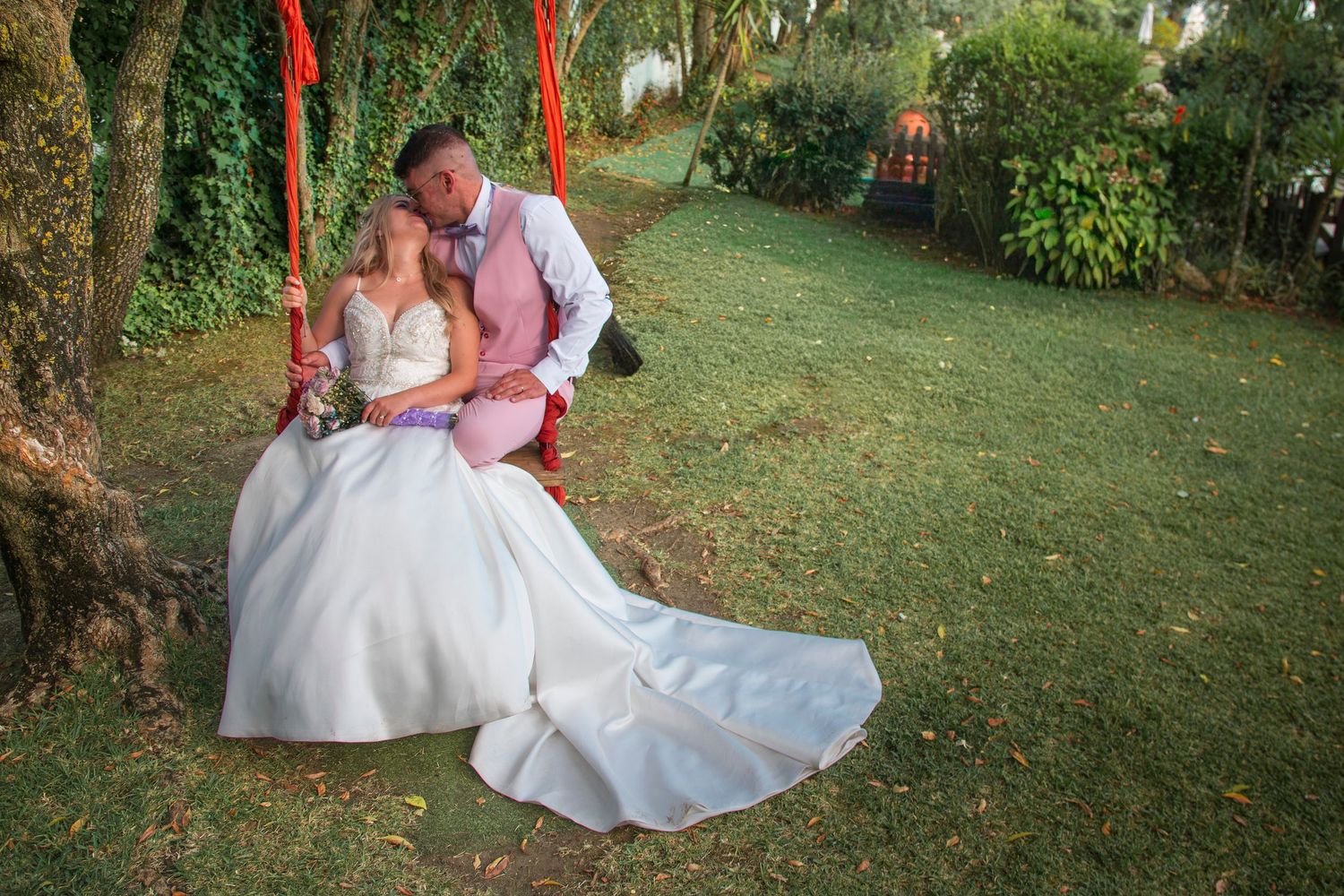 Bride in white gown and groom in pink suit share a romantic moment on a red swing in a lush green garden.